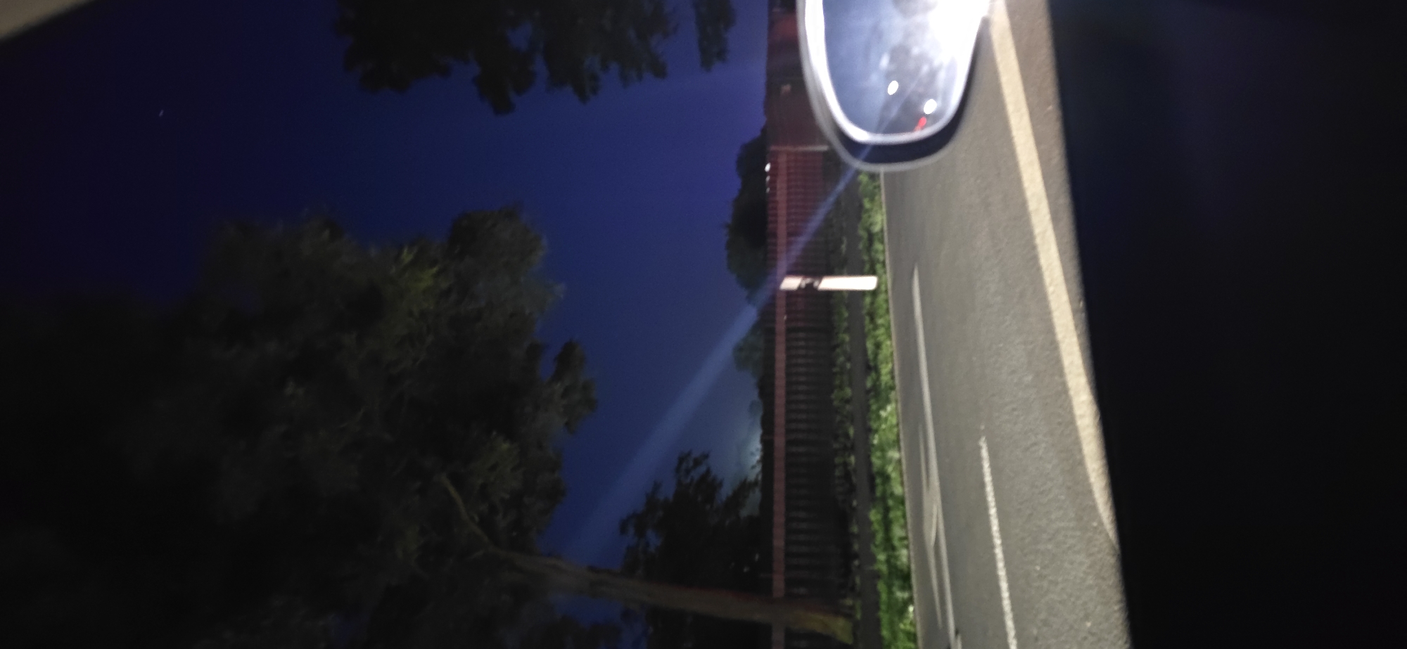 The image depicts a nighttime scene viewed from a vehicle, likely a car, driving along a road. The sky is a deep, dark blue, characteristic of twilight or a clear night. In the foreground, the right side of the image shows a car's side mirror, reflecting lights from the vehicle's interior or the surrounding environment. The road surface is visible, with white lane markings indicating it's a paved road. To the left and extending into the background, trees with dark foliage frame the scene. A light beam or ray cuts across the sky from the left towards the center, suggesting a light source from beyond the frame or a spotlight. Behind the trees and a wooden fence, a subtle glow of light suggests distant city lights or buildings. The overall impression is that of a drive through a semi-urban or rural area at night, possibly near Neustadt-Glewe, Germany, given the context provided.