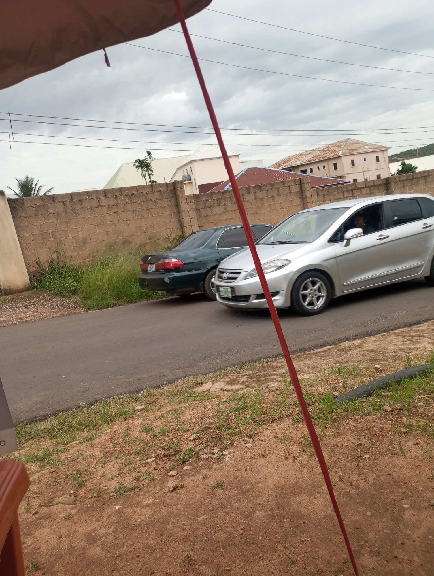 In Jos, Nigeria, a view from under a canopy shows two cars parked on a paved road. A dark green sedan is parked on the left, and a silver minivan is on the right. Behind the cars, a light brown brick wall runs horizontally across the frame, with overgrown green grass and plants growing in front of it. Beyond the wall, buildings with various roof colors, including red and gray, are visible, along with power lines against a cloudy sky. The foreground features a dirt and gravel path with sparse green grass.