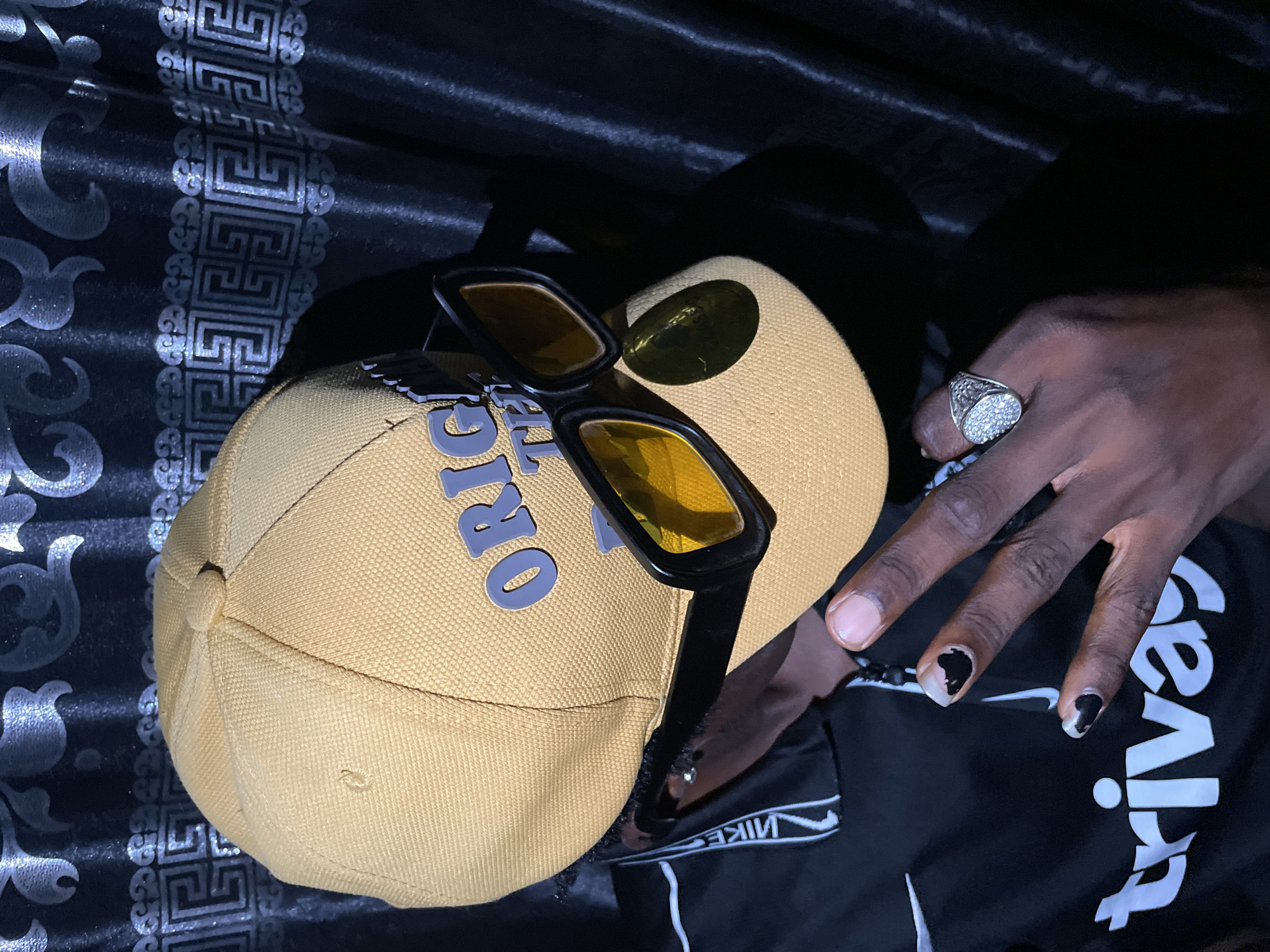 The image depicts a tan baseball cap with the word "ORIGINAL" printed in gray on the front. The cap sits on a black fabric with a repeating geometric pattern in silver. A pair of black-framed sunglasses with yellow lenses rests on the cap. A person's hand is visible, adorned with a large silver ring on the ring finger. The fingernails are painted with a black and white design. Part of a black Nike tracksuit is also visible. The location, Lokoja, Nigeria, cannot be verified from the image.