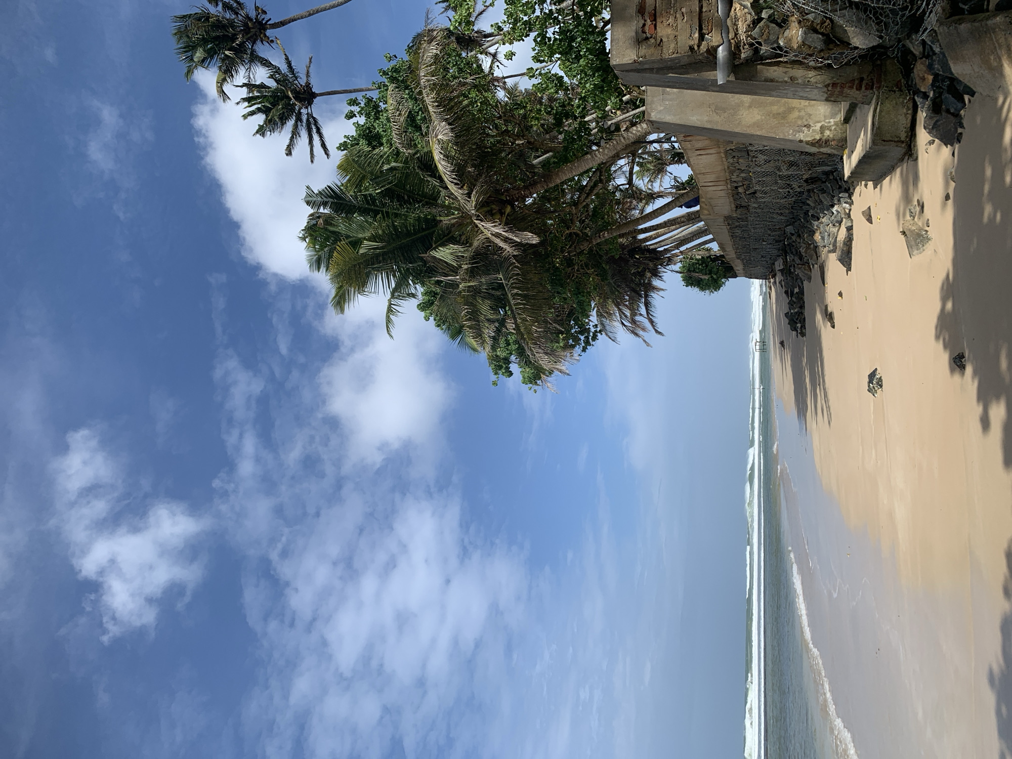 The image depicts a scenic view of Kathaluwa beach in Sri Lanka. The foreground is dominated by a sandy beach with scattered rocks and debris, leading to the ocean. The ocean is calm with gentle waves rolling onto the shore. In the background, a line of palm trees stands tall against a clear blue sky with scattered white clouds. A damaged concrete structure, possibly a wall or part of a building, is visible on the right side of the image, adorned with stones and wire mesh. The overall scene conveys a sense of natural beauty and tranquility.