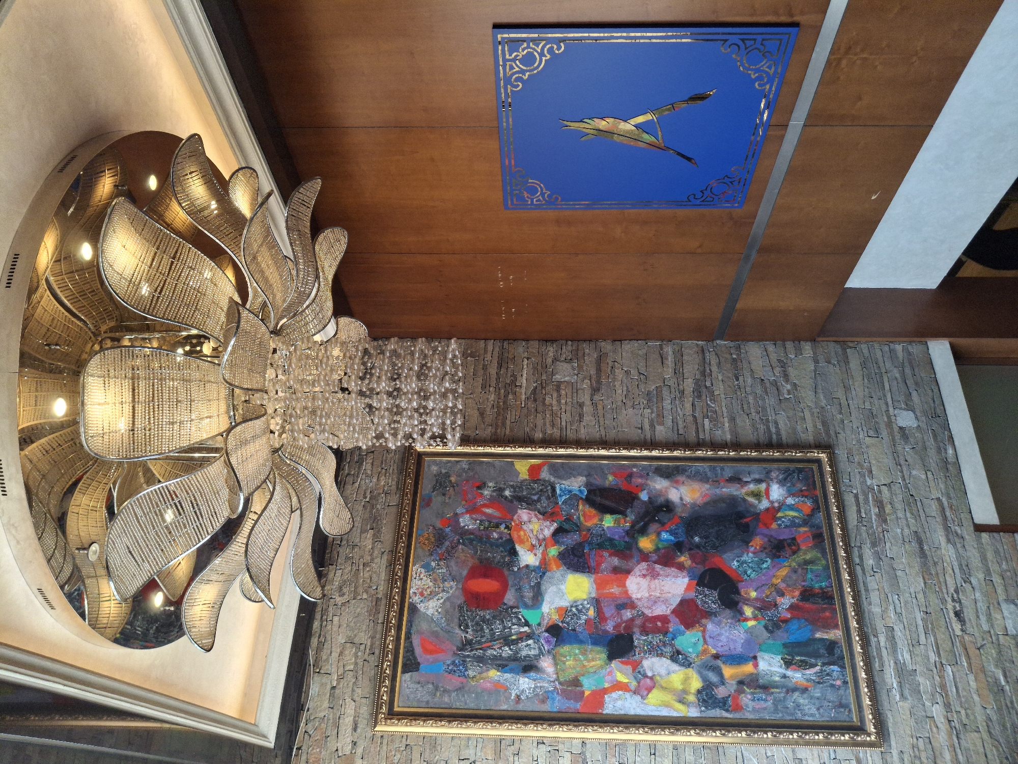 This media file captures a lavish interior space, primarily focusing on decorative architectural and artistic elements. The image is composed from a low-angle perspective, looking upwards at a wall and ceiling.
On the left, a large, ornate chandelier dominates the view. It features a multi-layered design with numerous sparkling, crystal-like beads forming cascading strands and curvilinear, metallic, leaf-like structures that reflect light. The chandelier emits a warm, bright glow, and its reflection is visible on a curved, polished surface behind it, enhancing its grandeur.
To the right of the chandelier and on the wall, a vibrant abstract painting is prominently displayed within an elaborate gold frame. The artwork is characterized by a dynamic interplay of bright colors such as red, yellow, blue, green, and purple, alongside darker shades of black and grey. It appears to be a mixed-media piece, possibly incorporating collage elements, creating a textured and complex visual.
Above the painting and extending across the ceiling are smooth, polished wooden panels, giving a rich and warm tone to the space. Integrated into these panels is a rectangular blue decorative panel. This panel features a stylized, gold-outlined bird-like figure in flight, with subtle gold patterns adorning its borders, suggesting an elegant, possibly East Asian or Art Deco influence.
The wall beneath the ceiling and behind the painting is clad in textured stone or stacked tiles, providing a natural, rugged contrast to the smooth wood and intricate decor. The lighting in the room is artificial, emanating primarily from the chandelier, and suggests an indoor setting, likely a hotel lobby, an upscale restaurant, or a grand residence, designed with a blend of modern luxury and traditional craftsmanship. There is no visible text.
