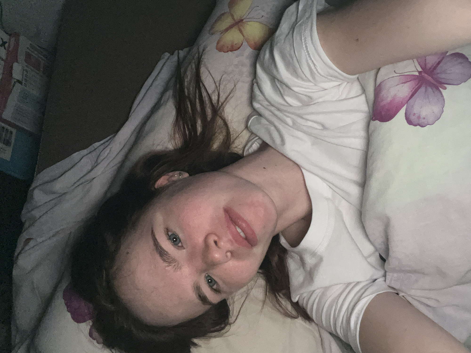 A selfie of a young woman in a white t-shirt lying on a bed. The bed has a pillow with a purple butterfly pattern and another with a yellow butterfly. The woman has fair skin, blue eyes, and brown hair. Part of a cardboard box is visible to the left of the bed. The location is possibly Chaikovsky, Russia.