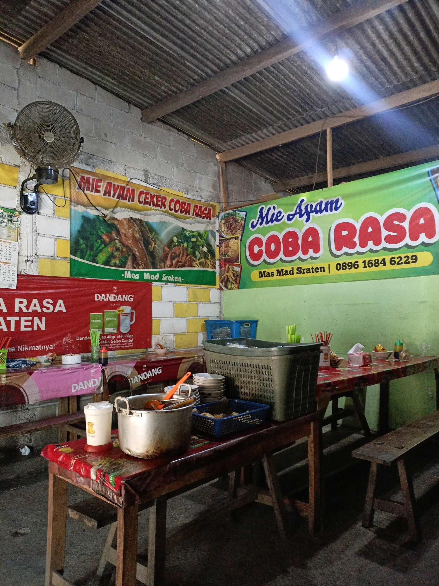 The image depicts the interior of a modest eatery, likely a noodle shop, in Sraten, Indonesia. The space appears utilitarian, with a corrugated metal roof supported by wooden beams. The walls are constructed of cinder blocks. A vintage wall-mounted fan is visible on the left side of the frame, and a bright fluorescent light illuminates the scene from the ceiling.
Prominently displayed are two large banners. The upper banner, predominantly yellow, features the text "MIE AYAM CEKER COBA RASA" with an image of chicken noodles. Below it, a red banner reads "RASA" and "DANDANG" with images of small boxes and a mug, suggesting a product advertisement. The second, larger green banner further down reads "Mie Ayam COBA RASA" with a phone number and the name "Mas Sraten".
Several tables are set up for dining. The tables are covered with patterned cloths. On one table, a large metal pot with a lid sits alongside a plastic cup and some utensils. Another table holds stacked plates, a basket filled with utensils, and small bowls of condiments. Wooden benches are positioned along the sides, indicating a casual dining setup. The floor appears to be concrete. The overall impression is of a local, no-frills establishment serving Indonesian noodle dishes.