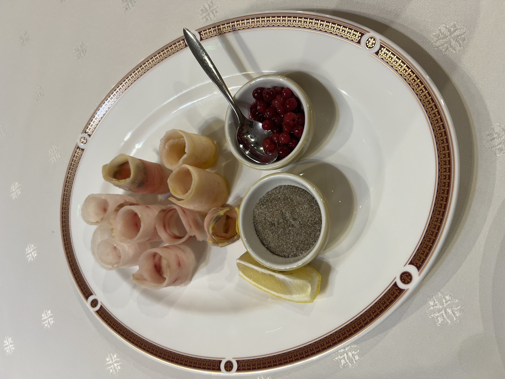 A white porcelain plate with a decorative gold and maroon rim sits on a white tablecloth with a subtle embossed pattern. The plate holds a platter of appetizer-sized rolled food items, appearing to be thinly sliced ingredients rolled into cylinders. Some of the rolls have a pinkish hue, suggesting a meat or fish component, while others are paler, possibly indicating a vegetable or cheese filling. Arranged around the rolled items are three small accompaniments: a wedge of bright yellow lemon, a small white bowl filled with a dark, granular substance resembling coarse ground pepper or spices, and another small white bowl containing a generous portion of small, red berries, possibly cranberries or lingonberries. A small silver spoon rests in the bowl of berries. The lighting suggests an indoor setting. The overall presentation is that of a carefully arranged appetizer or tasting plate.