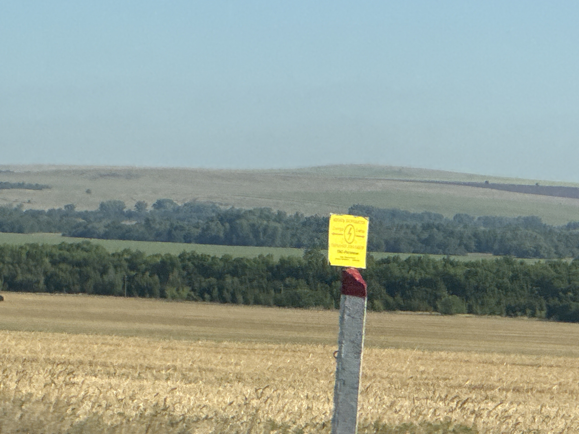 The image shows a rural landscape under a clear blue sky. In the foreground, a marker post with a yellow warning sign is visible. The sign has a black lightning bolt symbol and text in what appears to be Russian. The post is cylindrical and has a red and white band near the top. Behind the post, a field of dry, golden grass or wheat extends into the middle ground. Further back, a dense line of green trees forms a treeline. In the background, rolling hills covered in green vegetation are visible, with a hazy, light-colored sky above. The overall scene depicts an outdoor environment, possibly agricultural land, with a warning marker indicating some form of infrastructure. The image does not show any people, animals, or specific urban elements that would place it directly within the city of Gamaleevka-1, Russia, though the sign might suggest a location in Russia.