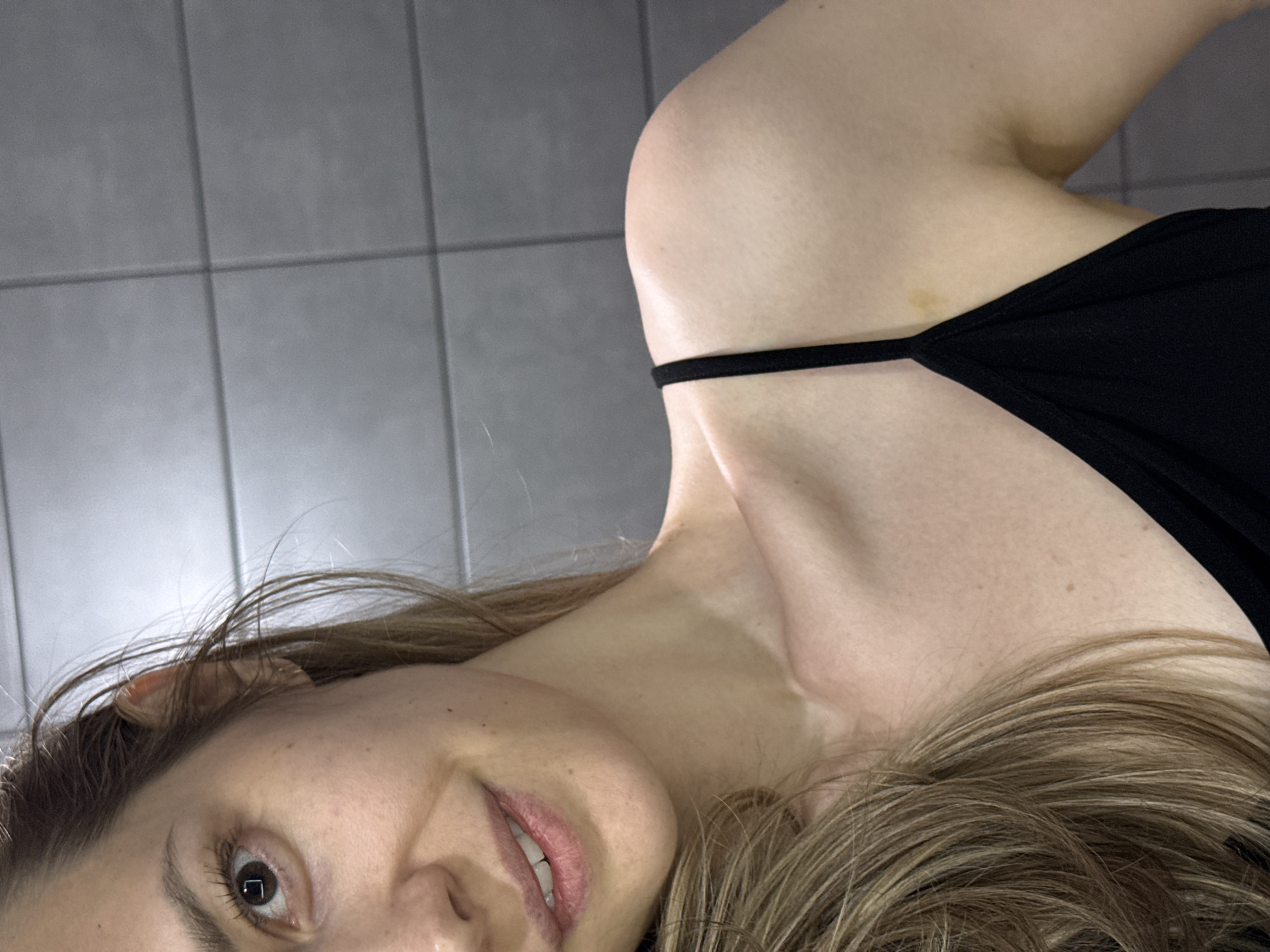 A woman is positioned close to the camera, angled towards the top-right. She is wearing a black top with a thin strap, and her blonde hair is visible. Her face is slightly turned towards the camera, showing one eye, part of her nose, and a partial view of her mouth with teeth. The background features grey, square tiles. There is no indication that this image was taken in Chaikovsky, Russia.