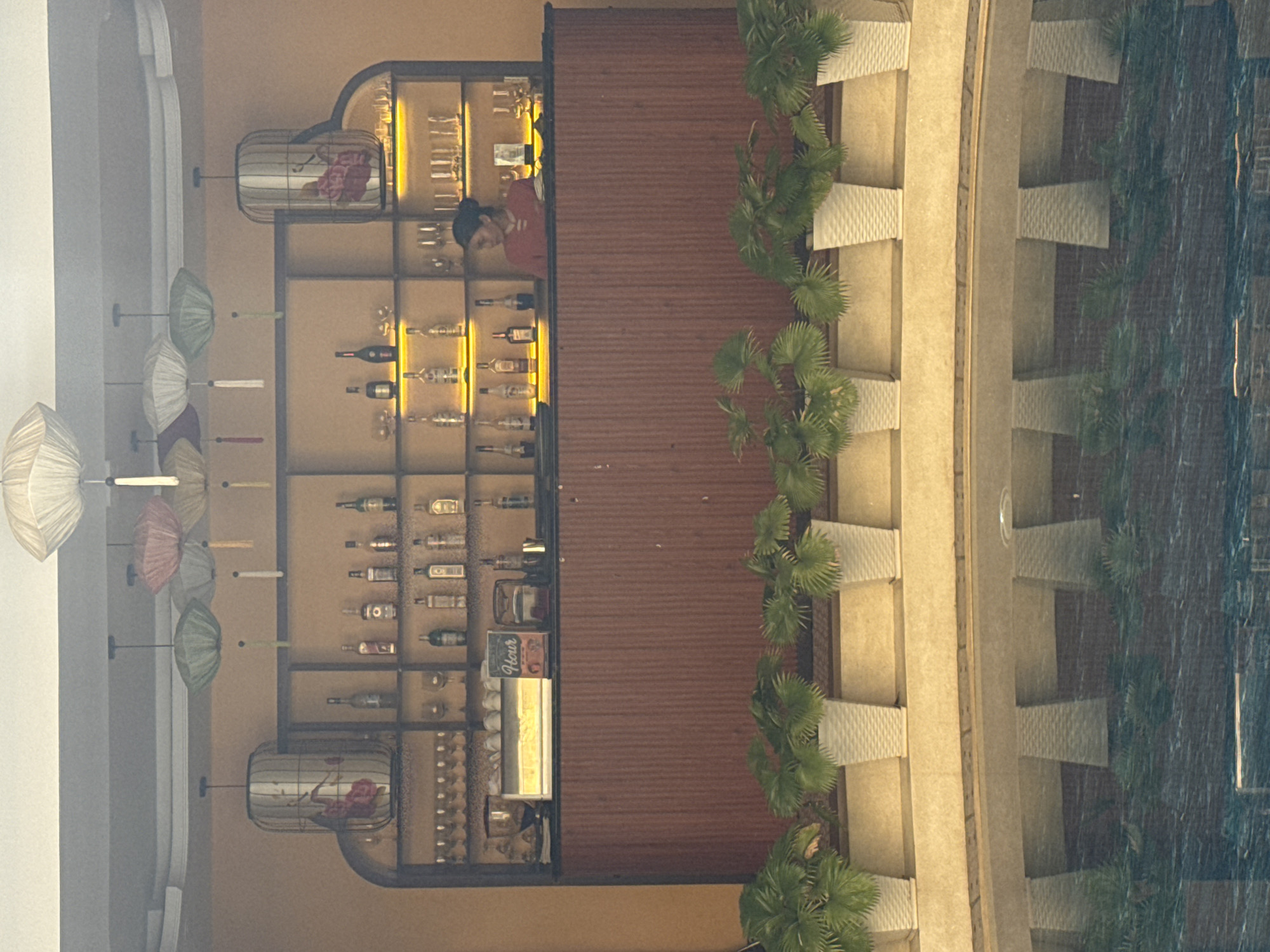 Captured from above, the image shows an outdoor bar area next to a body of water, likely a pool. The bar features shelves stocked with alcohol and glassware. A person in a red shirt is visible behind the bar. Small palm trees line the edge of the bar area. A row of decorative umbrellas is displayed against the wall. This scene is in the city of Đà Nẵng, Vietnam.