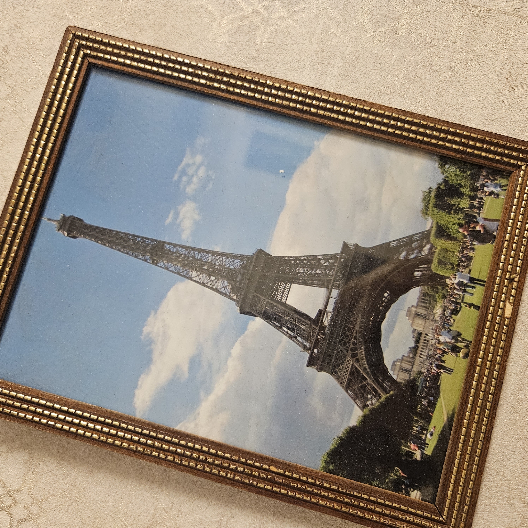 The image is a framed photograph of the Eiffel Tower in Paris, France. The photograph is tilted and appears to be placed on a textured, light-colored surface. The Eiffel Tower is the central subject, a tall iron lattice tower, seen against a blue sky with scattered white clouds. At the base of the tower, there is a grassy area with numerous people gathered. Some individuals are standing in groups, while others are walking or appear to be taking photos. Trees are visible on the left and right sides of the frame, and in the background, there are buildings with classical architecture. The photograph is framed by a decorative, golden-brown frame with a beaded texture. The image does not depict any location in the city of Chelyabinsk, Russia.