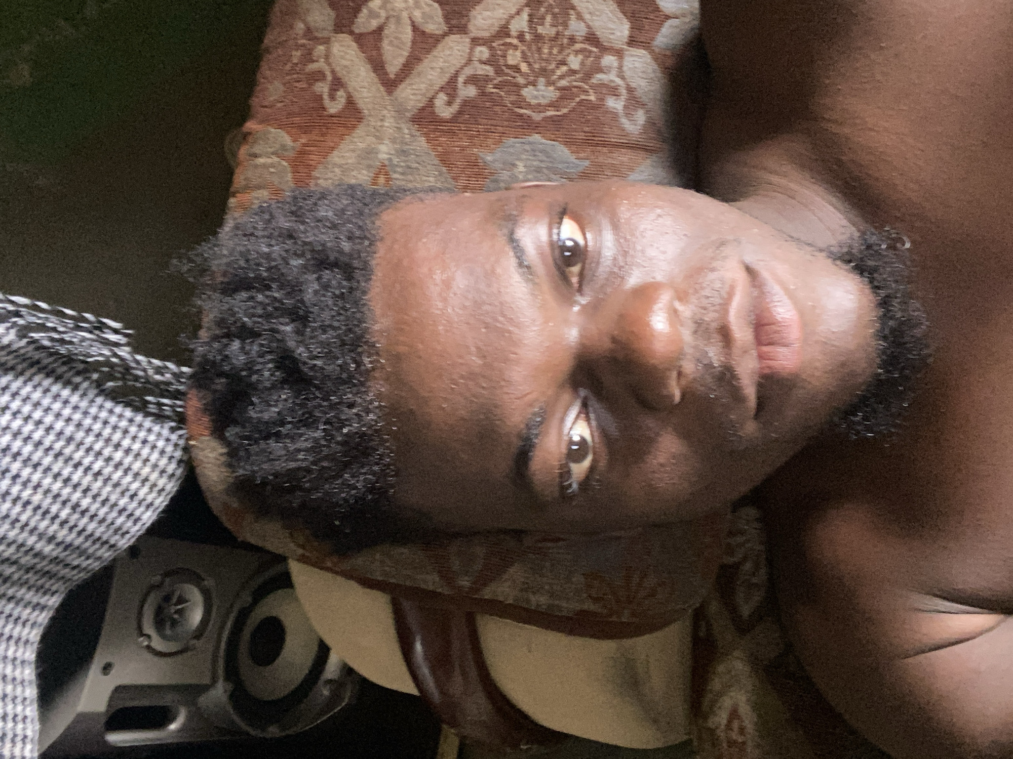 A portrait of a Black man with short, dark curly hair and a dark beard is captured in a close-up shot. He is lying on a patterned fabric surface. His skin appears to be slightly shiny. To the lower left of the frame, parts of a black and white checkered fabric are visible along with a metal object containing screws. The city and location is unknown.