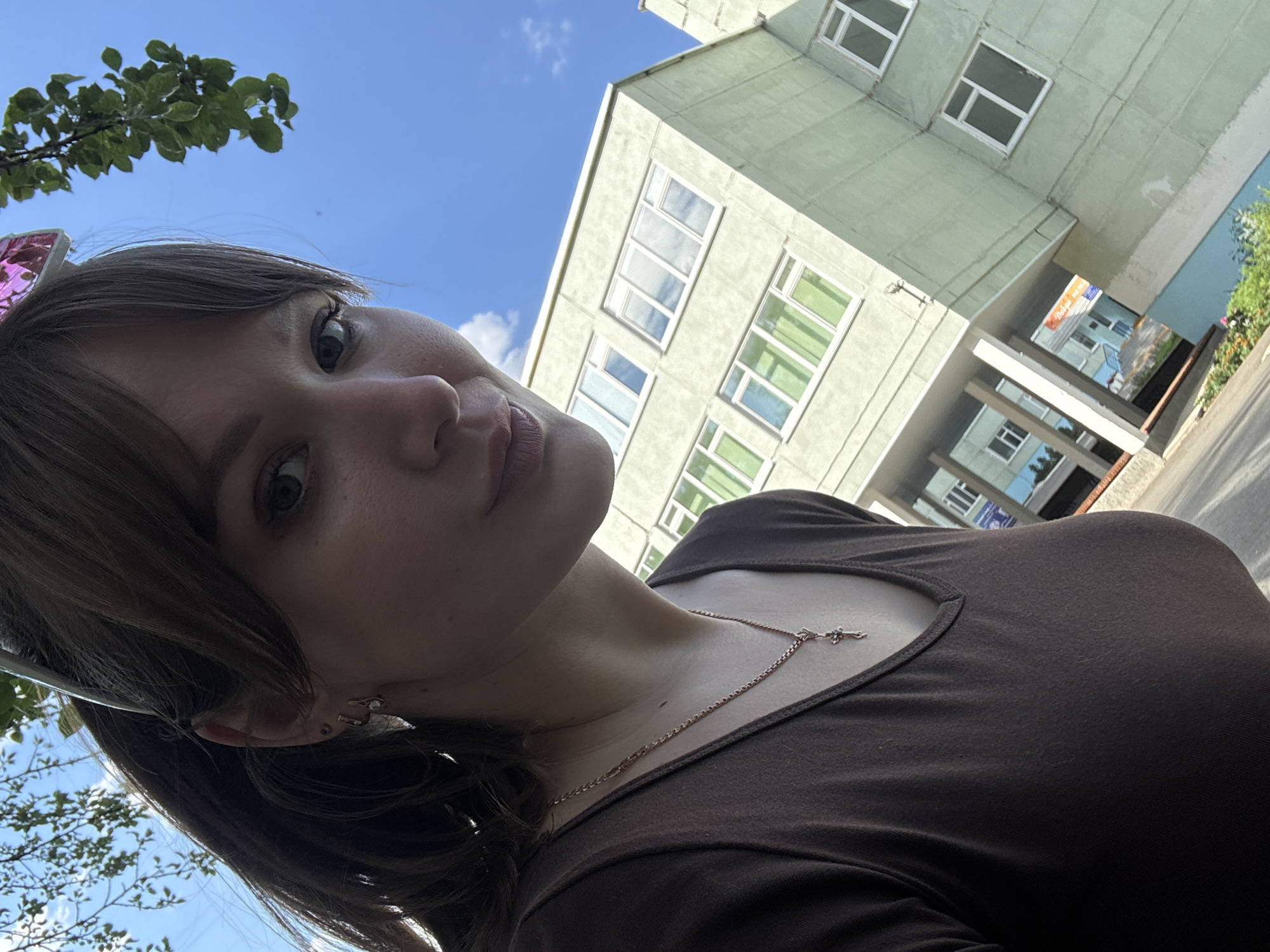 A young woman with brown hair, wearing a brown top and a rose gold cross necklace, is taking a selfie outdoors. She is wearing pink bunny ears with glitter. The background shows a pale green, multi-story building with many windows. To the left of the frame, there is a tree branch with green leaves against a bright blue sky with some white clouds. The lighting suggests it is daytime, likely sunny. The woman's expression is neutral, with her lips slightly pursed. Her left ear is visible, adorned with two small earrings. A street or walkway is visible in the lower right portion of the image. The scene appears to be in an urban or suburban residential area.