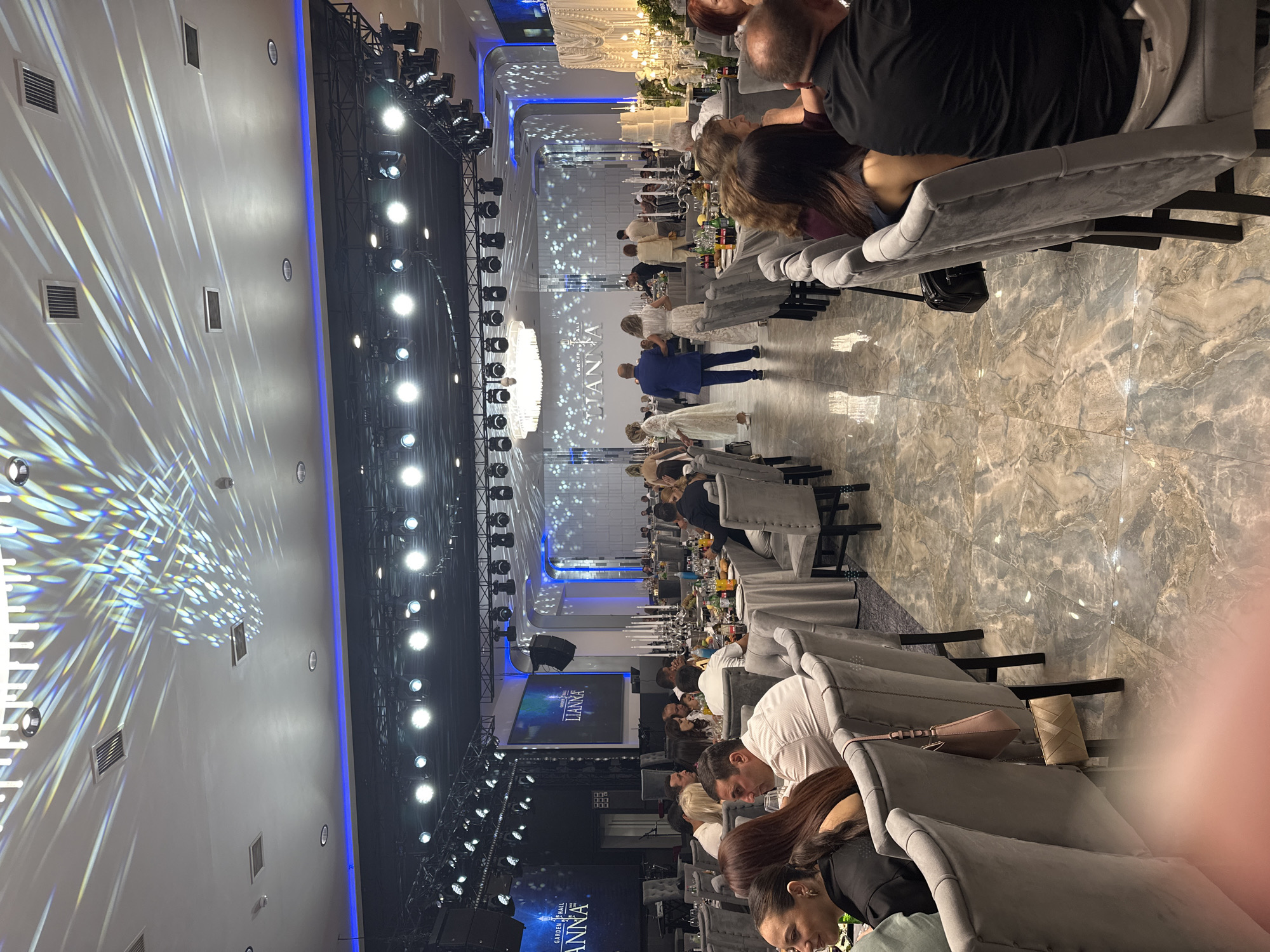 The image depicts a formal indoor event, likely a wedding reception, taking place in the Garden Hall in Parakar, Armenia. The hall is elaborately decorated with a large chandelier, spotlights, and illuminated screens displaying the event's name, "LIANNA." Guests are seated at round tables covered with gray tablecloths, with elaborate centerpieces featuring candles. Several people are visible in the foreground and midground, engaged in conversation or observing the surroundings. In the background, a bridal couple, dressed in white, walks with other attendees, suggesting a key moment in the event. The flooring is a reflective marble-like tile. The overall atmosphere is one of celebration and elegance.