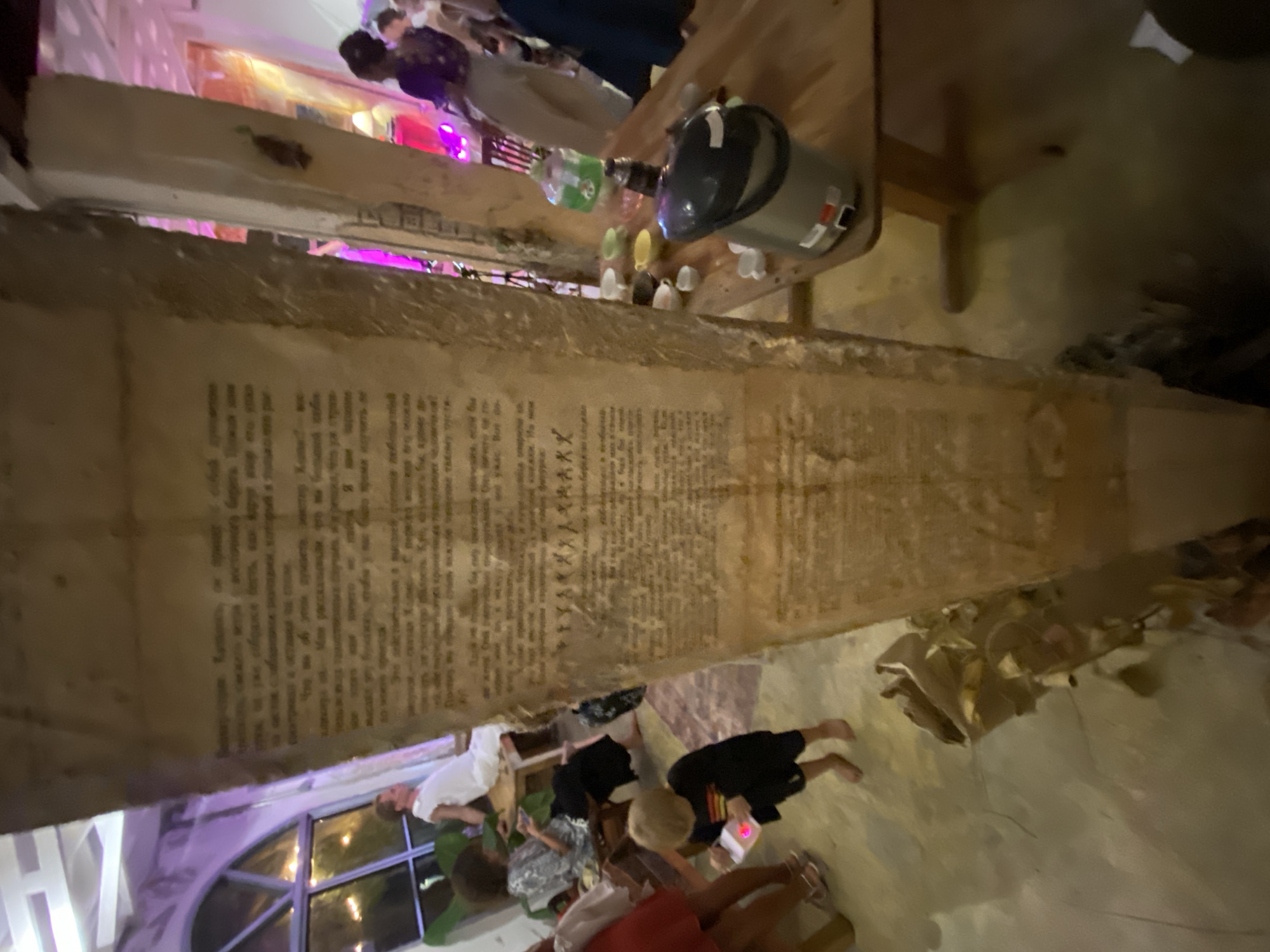 In a dimly lit indoor setting, possibly a party or event, several people are gathered. The image is oriented vertically. In the foreground, a large, rough-hewn pillar-like structure is prominently displayed, covered with what appears to be old text or a document. The text is too blurry to read specifically, but it's dense and arranged in columns.
Above and behind the pillar, a wooden table holds several objects, including a dark-colored portable dispenser or jug with a handle, and several small, colorful bottles or cups. Some people are visible in the background, appearing to interact or observe. One person is wearing a white shirt, another a dark shirt. There are also hints of colored lighting, particularly purple and pink, suggesting a festive atmosphere.
In the lower portion of the image, more people are visible, some seated and some standing, also appearing to be part of the gathering. One person is wearing a black outfit, and another is in a red top. The floor appears to be a light-colored, possibly concrete or tiled surface. The overall mood seems casual and social, with the illuminated text on the pillar being a central, intriguing element of the scene.