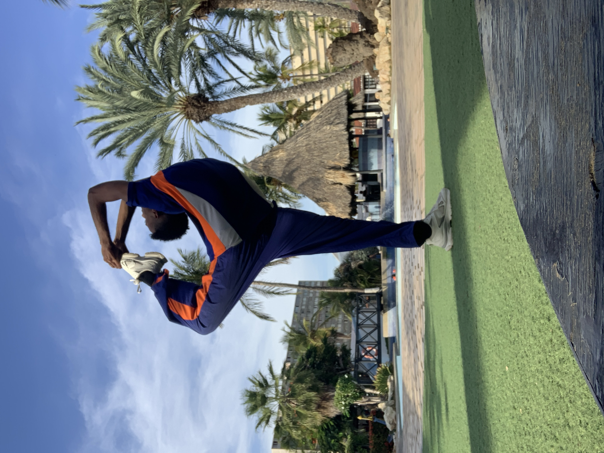 This outdoor media file captures a person performing an athletic pose in a tropical resort setting, likely in Porlamar, Venezuela.
**Subjects:** The central subject is a person, appearing to be male, with dark skin and short dark hair, dressed in a blue and orange athletic tracksuit and white athletic shoes. Surrounding them are numerous tall palm trees, a multi-story building (possibly a hotel), and a small thatched-roof hut (palapa).
**Scene and Setting:** The scene is set in an outdoor area, indicative of a resort or leisure complex. The immediate foreground features a section of vibrant green artificial turf, bordered by a dark, weathered wooden plank or deck. Beyond the turf and trees, a glimpse of a swimming pool is visible. The sky above is clear and bright blue with scattered white clouds.
**Activity:** The person is actively engaged in a display of flexibility or a gymnastic movement. They are standing on one leg, with the other leg extended high behind them, nearly vertical. Both hands are clasped around the upward-pointing foot, pulling it further into the arched back pose. This action suggests a trained athlete or dancer.
**Notable Details:** The image is taken in bright daylight, indicated by the well-lit scene and clear sky, suggesting either morning or afternoon. The overall atmosphere is sunny and pleasant. The architecture (thatched hut, resort building) and abundant palm trees strongly suggest a tropical vacation destination. There is no visible text in the image.