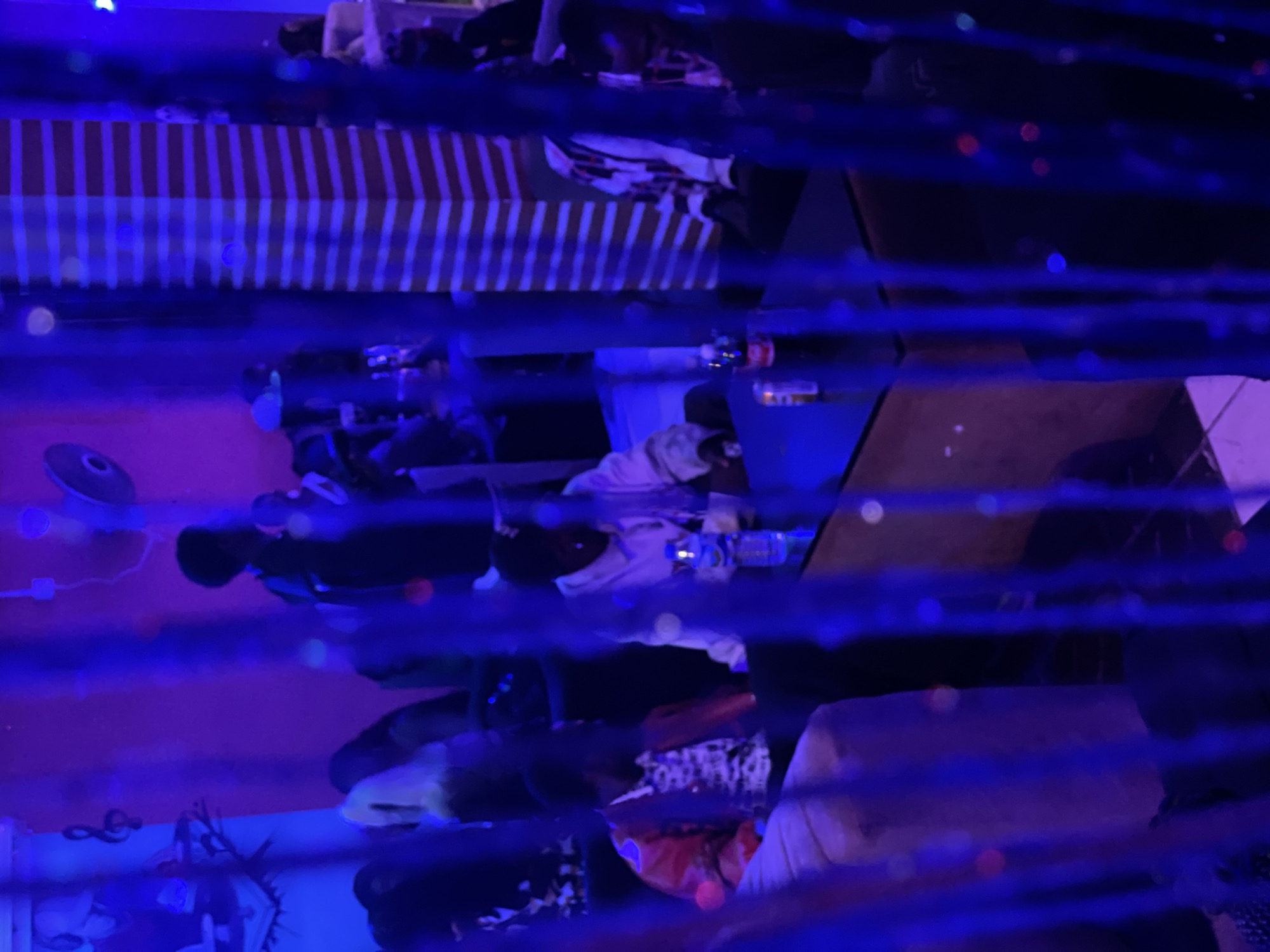 The image depicts an indoor scene at a dimly lit establishment, likely a bar or lounge, in Durumi, Nigeria. The ambiance is characterized by the pervasive blue and purple lighting, creating a nightclub-like atmosphere. In the foreground, blue beaded curtains partially obscure the view.
Several individuals are present, seated around tables. On the left, a person wearing a red jacket over a patterned shirt is seated in a beige armchair. To their right, another person in a white hoodie is sitting. A small table between them holds a bottle of water and a can.
Further into the scene, other patrons are visible, though their features are less distinct due to the lighting and distance. A striped pillar, with alternating red and white horizontal bands, stands prominently in the mid-ground. The walls appear to be painted in shades of pink and purple, with some decorative elements like a musical note and possibly artwork or posters on the left side. The floor is tiled in a mosaic pattern. The overall impression is of a social gathering in a vibrant, albeit dimly illuminated, setting within Durumi.
