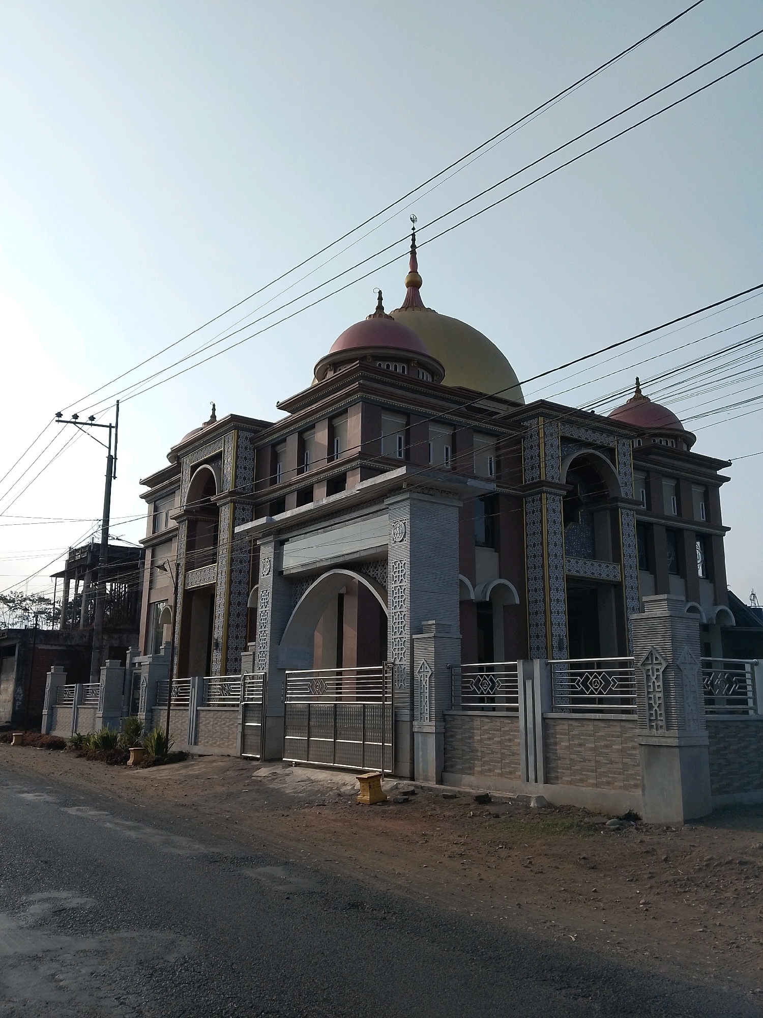 The image shows a mosque in the city of Wonokerto, Indonesia. The mosque is a multi-story building with several domes, the largest of which is golden. The building is constructed with brown and gray materials, and features decorative tiling around the arched entrances and windows. A metal fence with an intricate geometric pattern encloses the front of the property, and a large metal gate is visible. The mosque is located on a street with a paved surface that appears to be in fair condition, with some gravel and dirt visible. Electrical wires are strung across the sky above the building, supported by a utility pole on the left side of the image. There are no people visible in the scene. The sky is clear and light blue, suggesting it is daytime.