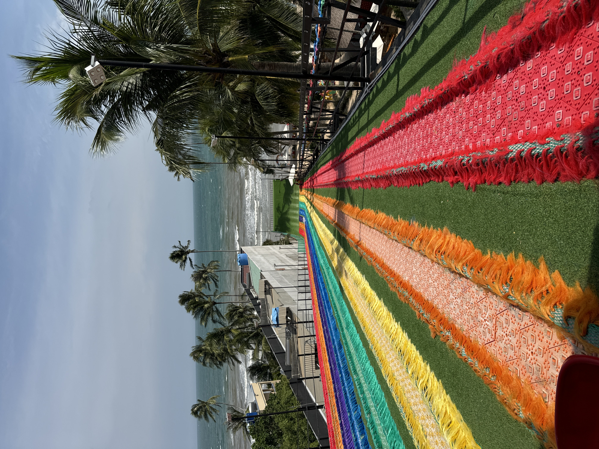 The image, taken in Mũi Né, Vietnam, depicts a vibrant outdoor scene with a focus on a colorful pathway leading towards the ocean. The pathway is constructed with rows of bright, fringed fabric in a rainbow gradient of colors, including red, orange, yellow, green, blue, and purple. These rows are laid over artificial green turf. The pathway appears to be part of an elevated structure, as indicated by the railing and wooden fencing on the right side of the frame.
In the background, the turquoise ocean meets a hazy sky with scattered clouds. Several palm trees, characteristic of a tropical coastal environment, are visible, some with lights attached to their trunks. A building with a sloped roof and some rooftop amenities, possibly part of a resort or hotel, is situated between the pathway and the ocean. The overall atmosphere suggests a resort or recreational area, likely a place designed for tourists to enjoy the scenery and perhaps engage in activities. There are no people clearly visible in the image.