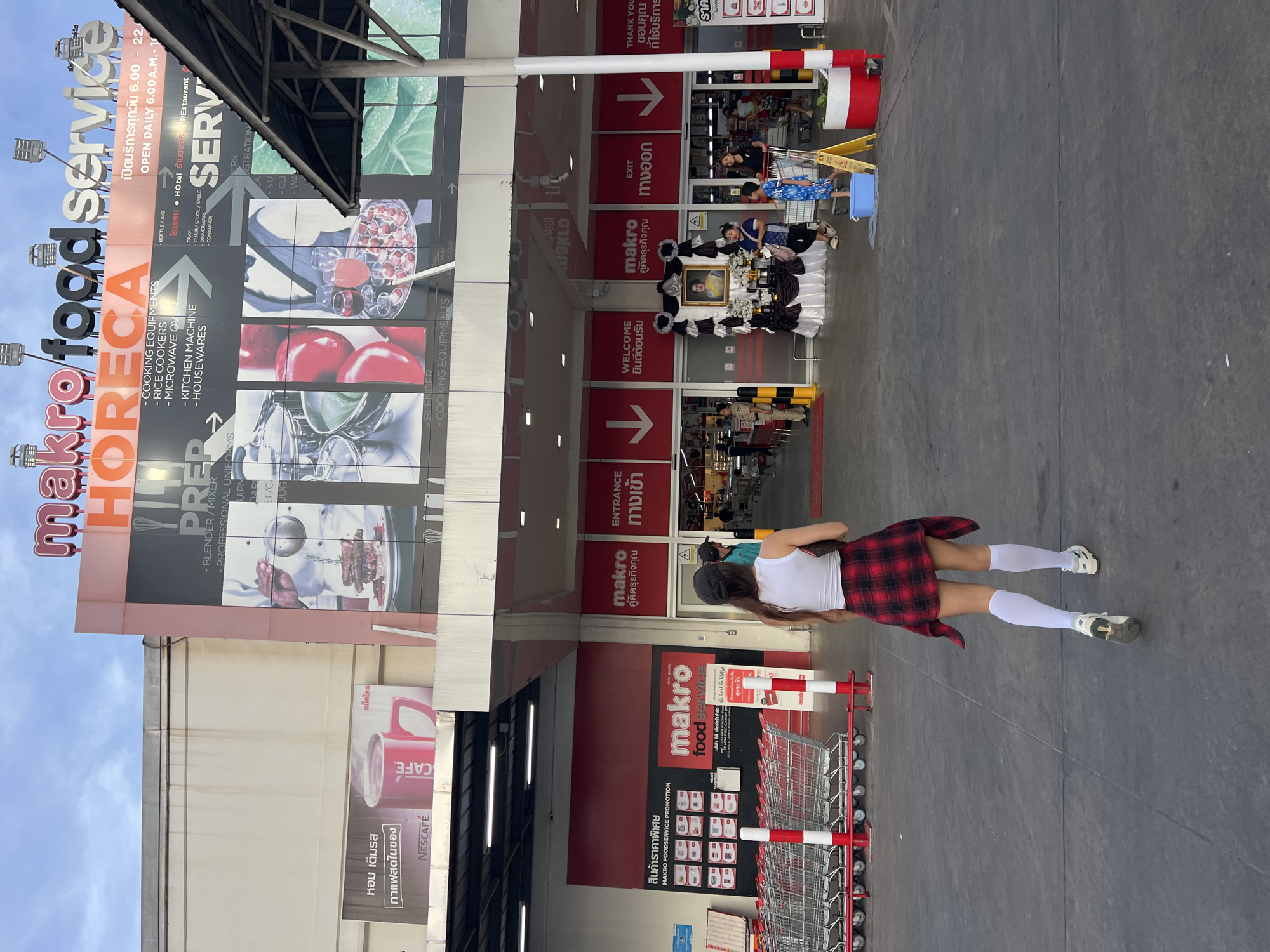 The image shows the exterior of a Makro Food Service store in Baan Bo Phut, Thailand. A woman with long hair, wearing a white tank top, a red and black plaid skirt, and white knee-high socks is walking towards the entrance. There are shopping carts lined up near the entrance. Banners and signs advertising various products and services are visible on the building's facade. A memorial table decorated with flowers and a portrait is set up near the entrance. Other customers can be seen inside the store.
