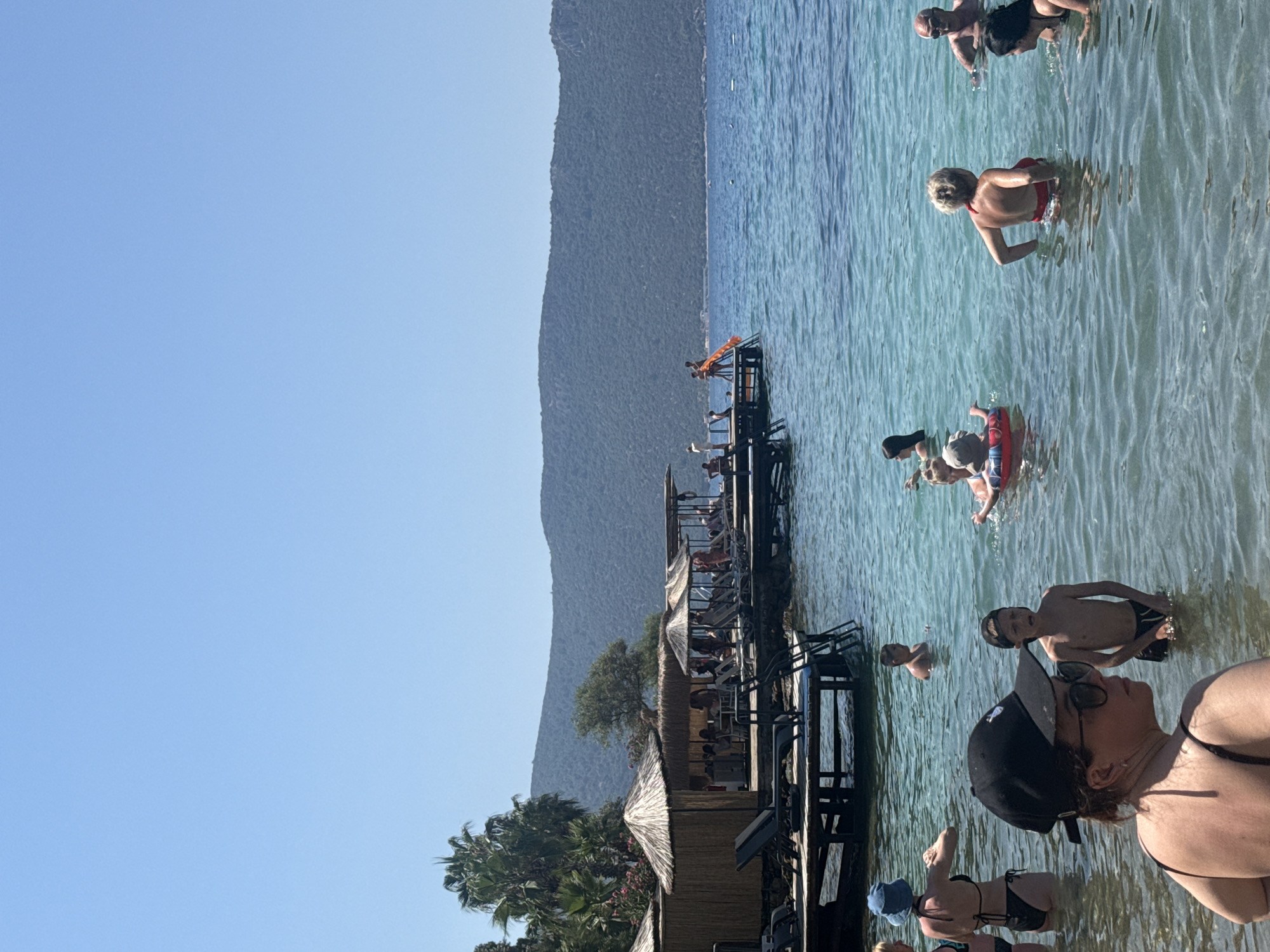 In Bodrum, Turkey, a vibrant scene unfolds at a beachfront establishment under a clear blue sky. A wooden pier extends into the turquoise waters, dotted with thatched-roof cabanas and sun loungers. People of various ages are enjoying a sunny day; some are wading in the shallows, while others are further out, swimming and playing. A young child is seen with a red inflatable ring, and another child stands in the water wearing a baseball cap. A woman in a black bikini and a blue sun hat faces away from the camera, her back to the sea. In the foreground, a woman wearing sunglasses and a black cap is partially visible, looking to her right. The backdrop features a lush green hillside that slopes down to the sea, creating a picturesque coastal landscape.