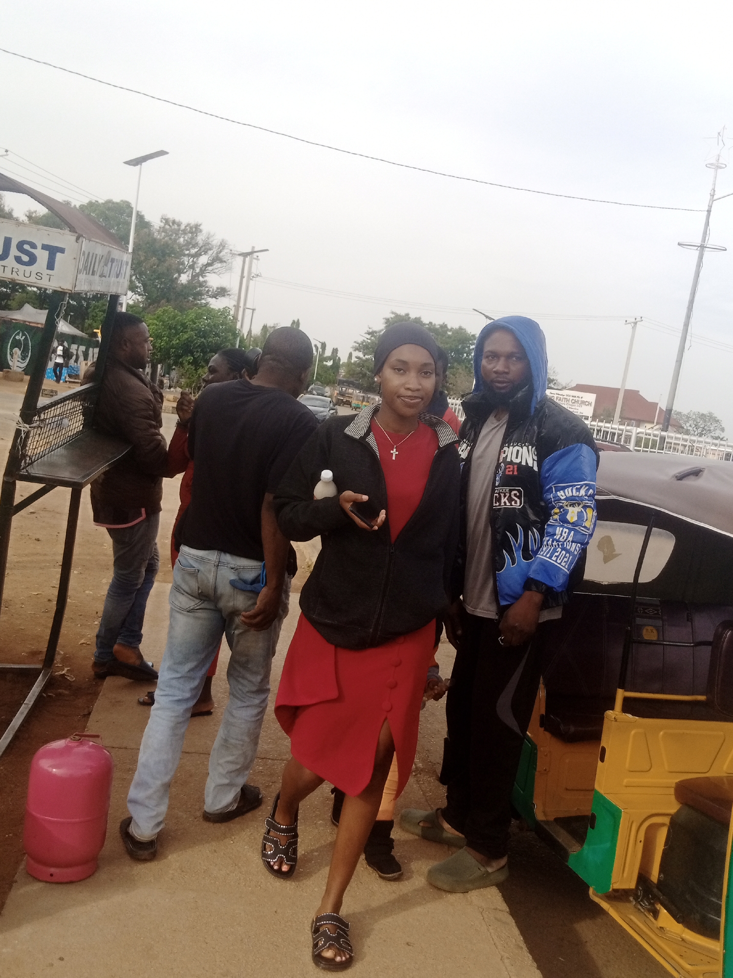 In Bar Jirgi Summa, Nigeria, a group of people stand on a paved street. The sky is overcast. A young woman in a red dress and a black jacket, adorned with a silver cross necklace, holds a white object. Beside her, a man wears a blue hooded jacket over a gray t-shirt and black pants. In the background, a man in a brown jacket and jeans stands near a metal stand. Another man in a black t-shirt and light blue jeans is visible. A pink gas cylinder rests on the ground. A yellow and green tricycle is parked to the right of the group. Various trees and buildings are visible in the distance.