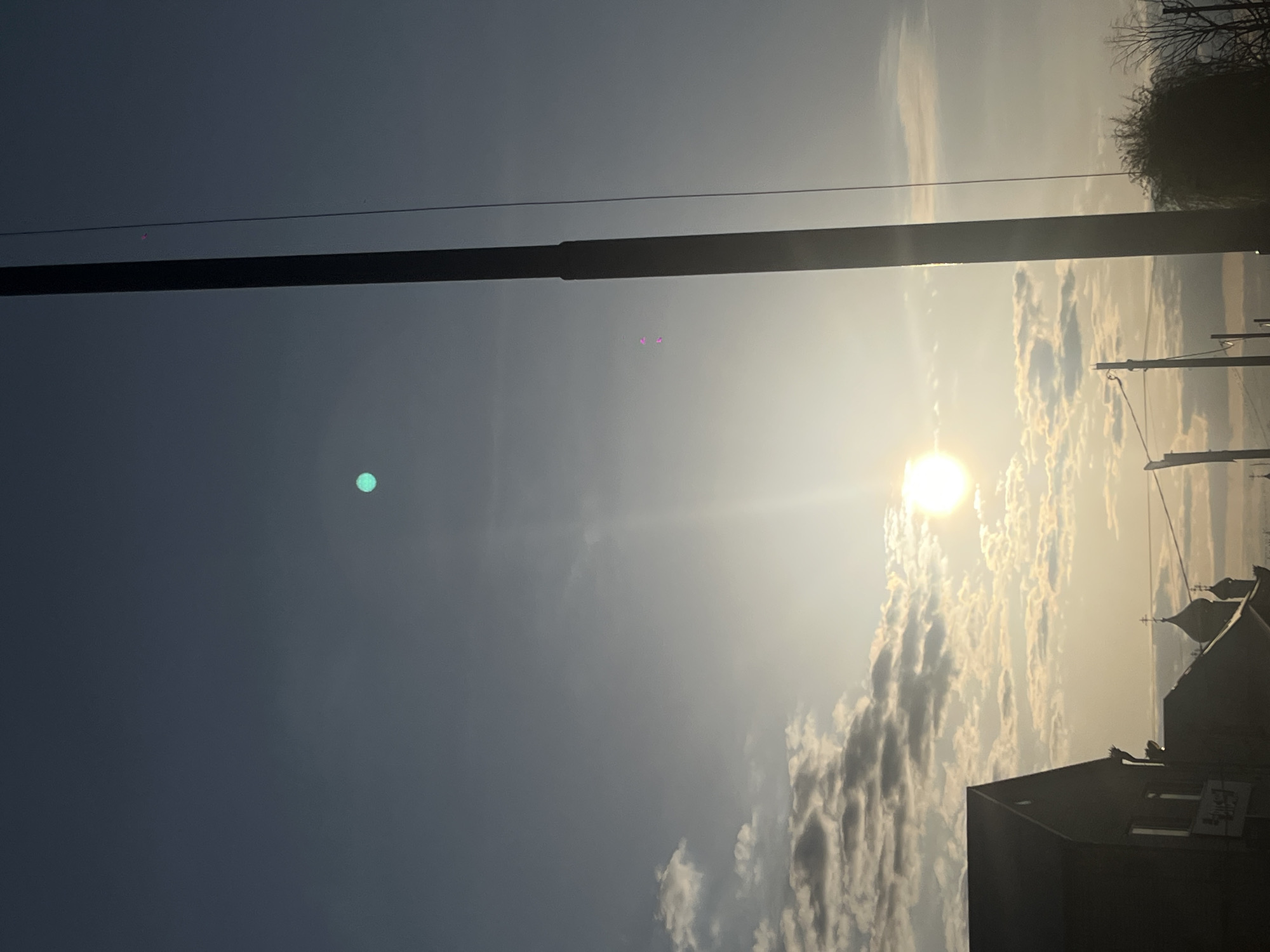 The image, when viewed correctly (rotated 90 degrees clockwise), displays an outdoor scene at either sunrise or sunset. A very bright sun is positioned low in the sky towards the right, casting a warm, golden glow across scattered cumulus clouds and creating significant lens flare. The sky gradient shifts from a deep blue on the far left to a brilliant, hazy white and yellow around the sun.
Prominent man-made structures are visible. A thick, dark utility pole or pillar stands vertically on the left side of the frame. Towards the right, two thinner utility poles with multiple horizontal wires are silhouetted against the bright sky. Along the bottom edge, the dark silhouettes of buildings are discernible, including one with a distinctive pointed roof or dome. A dark, bare tree or bush is also silhouetted in the upper right corner.
Notable atmospheric and photographic details include a distinct turquoise/green circular lens flare in the darker blue sky on the left and several small magenta flares near the center-top, resulting from the direct sunlight. The scene is devoid of visible human activity, focusing instead on the interplay of natural light and urban elements. The architecture and utility infrastructure are consistent with an urban or semi-urban environment in the described location of Novopokrovskaya, Russia. No visible text is present.
