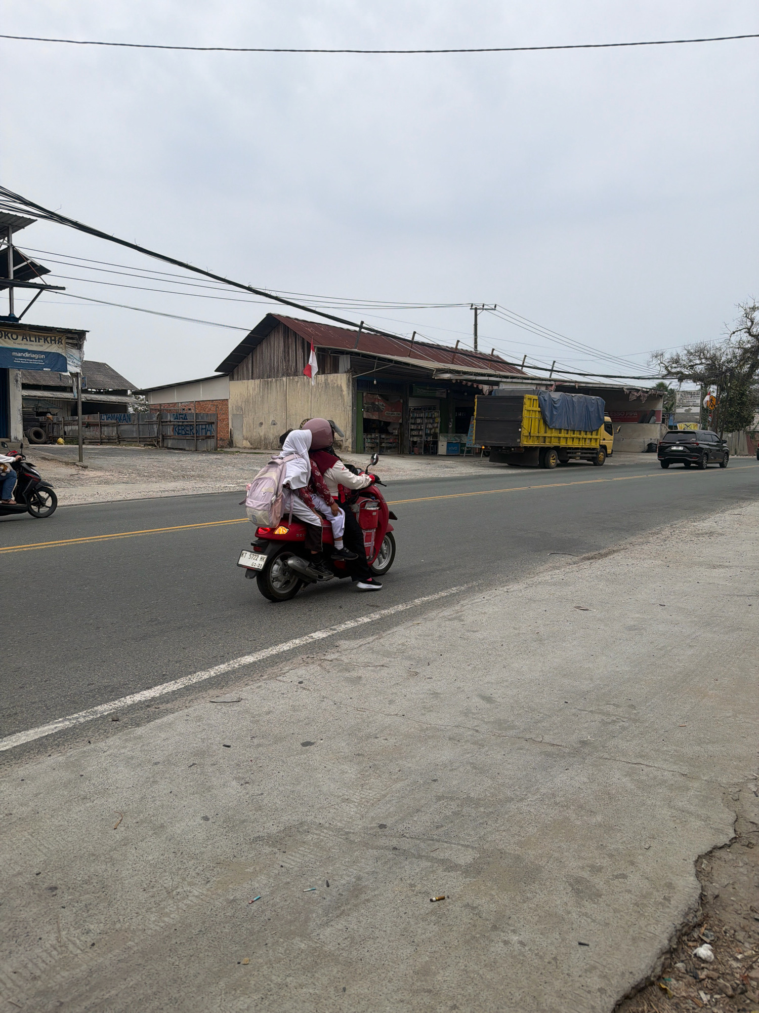 In Balikpapan, Indonesia, on a cloudy day, two individuals, likely a parent and child, are riding a red scooter on a paved road. The rear passenger, wearing a white hijab and a red jacket, carries a light pink backpack. The scooter has a license plate with the identifier "KT 5722 HK". A second scooter, ridden by one person, is visible in the background on the left. The roadside features various buildings, including a shop with a sign that reads "TOKO ALIFKHA" and a large yellow truck covered with a blue tarp. The scene depicts a typical street view in a developing urban environment.