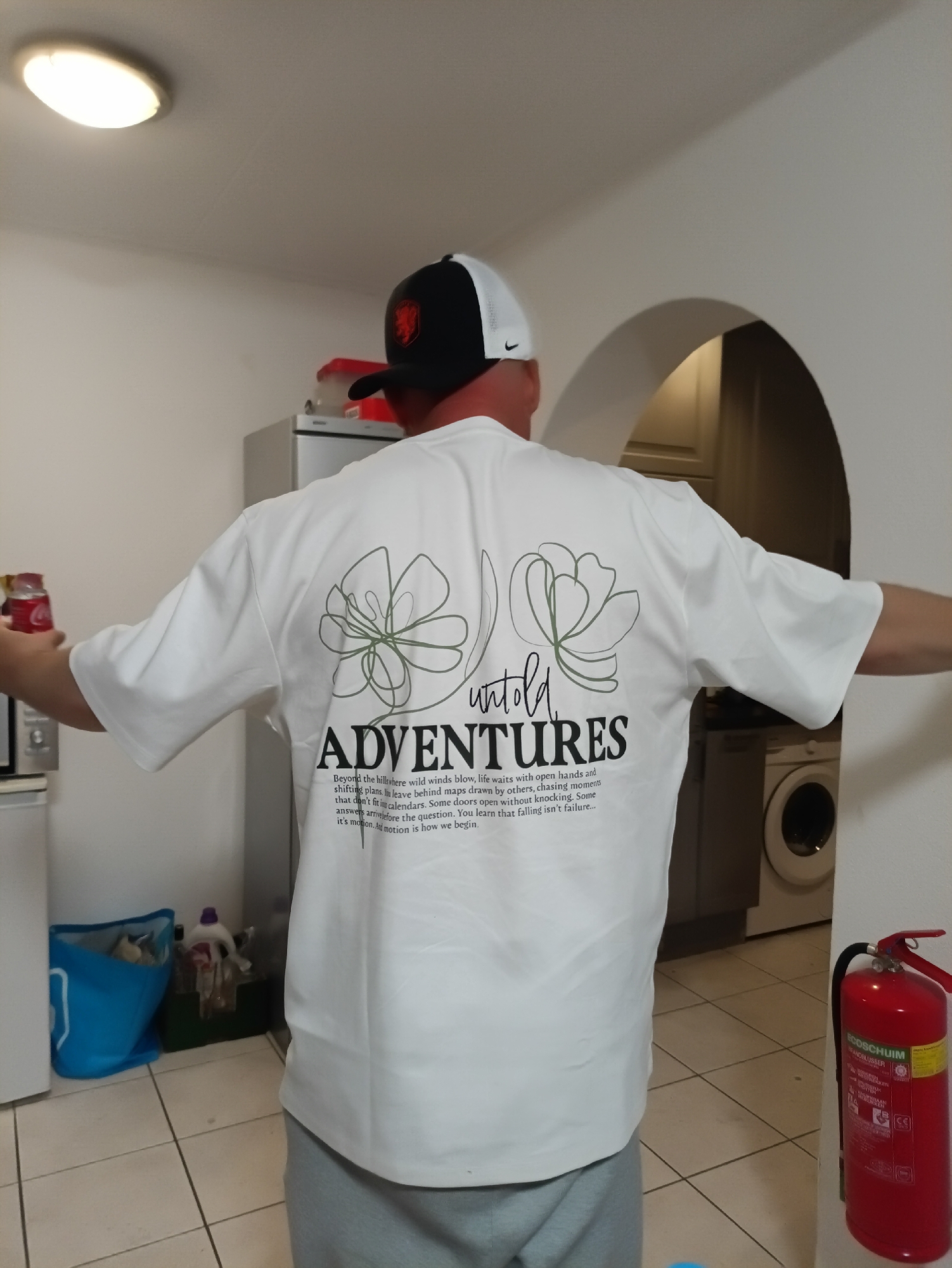 The image shows a person from behind, wearing a white t-shirt with a graphic design and text on the back. The design features two stylized green flowers and the words "untold ADVENTURES". Below the word "ADVENTURES", there is a block of text that reads: "Beyond the hills where wild winds blow, life waits with open hands and shifting plans. We leave behind maps drawn by others, chasing moments that don't fit into calendars. Some doors open without knocking. Some answers arrive before the question. You learn that falling isn't failure... it's motion. And motion is how we begin." The person is also wearing a black and white baseball cap.
The environment appears to be indoors, possibly a kitchen or utility area. Visible in the background are a refrigerator, a washing machine, a blue recycling bin, and a red fire extinguisher. The flooring is tiled.
The provided location of Zwartewaal, Netherlands, is not directly indicated by any visual cues within the image itself.