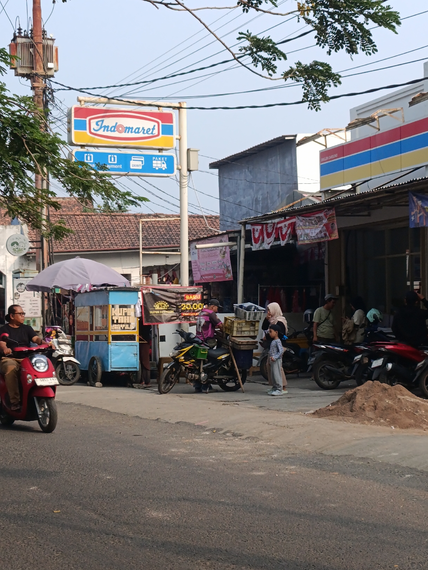 This is a street-level view of a commercial area in Cariu, Indonesia, on a clear day. Dominating the upper left of the frame is a utility pole with a transformer and a weathered Indomaret sign. Below the sign, a smaller blue sign indicates services like "visa payment," "payment," "DEBIT," and "PAKET." To the right, a building with a red, blue, and yellow striped awning suggests another commercial establishment.
In the foreground, a paved road is visible with a red scooter parked on the left, ridden by a man in a black shirt. A blue food cart with a light purple umbrella is situated on the sidewalk. This cart displays text that appears to say "KUPA TAHU" and has colorful graphics. Several motorbikes are parked behind the food cart and in front of the buildings.
People are present in the scene: a man on the red scooter, a woman in a beige hijab and a child in a striped shirt standing near the food cart, and several individuals near the buildings, some of whom appear to be customers or staff. The environment is urban, with a mix of buildings, overhead wires, and trees. A pile of sand is present on the right side of the sidewalk. The overall impression is of a bustling local marketplace.