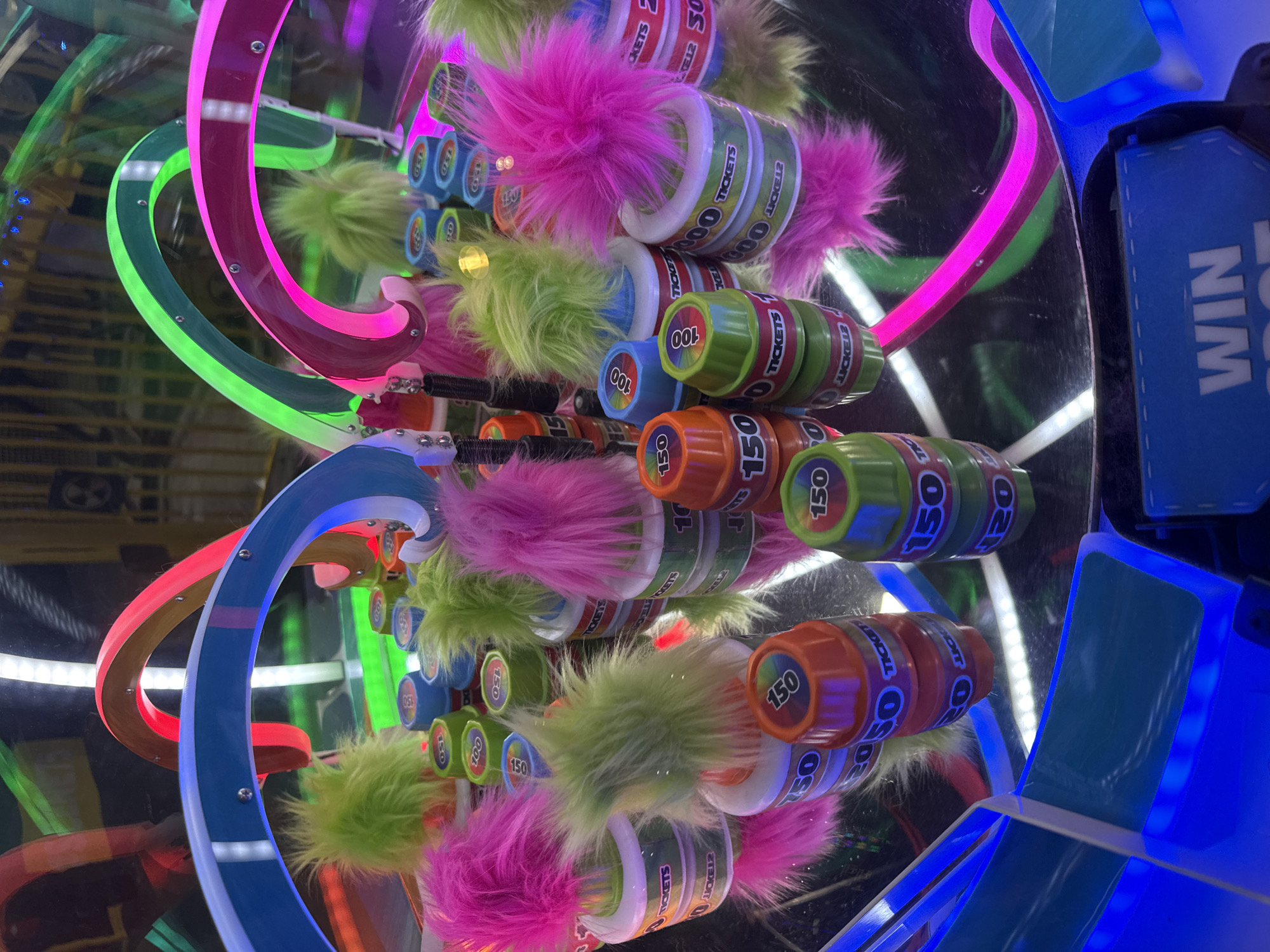 The image shows an arcade game in a reflective, mirrored enclosure, with brightly colored circular tracks in shades of blue, pink, red, and green illuminated by white LED lights. Cylindrical prize capsules are stacked within the game. Some of the capsules are adorned with colorful fluffy pom-poms (pink or green). The capsules have text that reads "TICKETS" and values such as "100" or "150". The game's control panel is visible on the right side, illuminated with blue light and features the word "WIN". There is no indication of specific location in Columbus, Ohio.