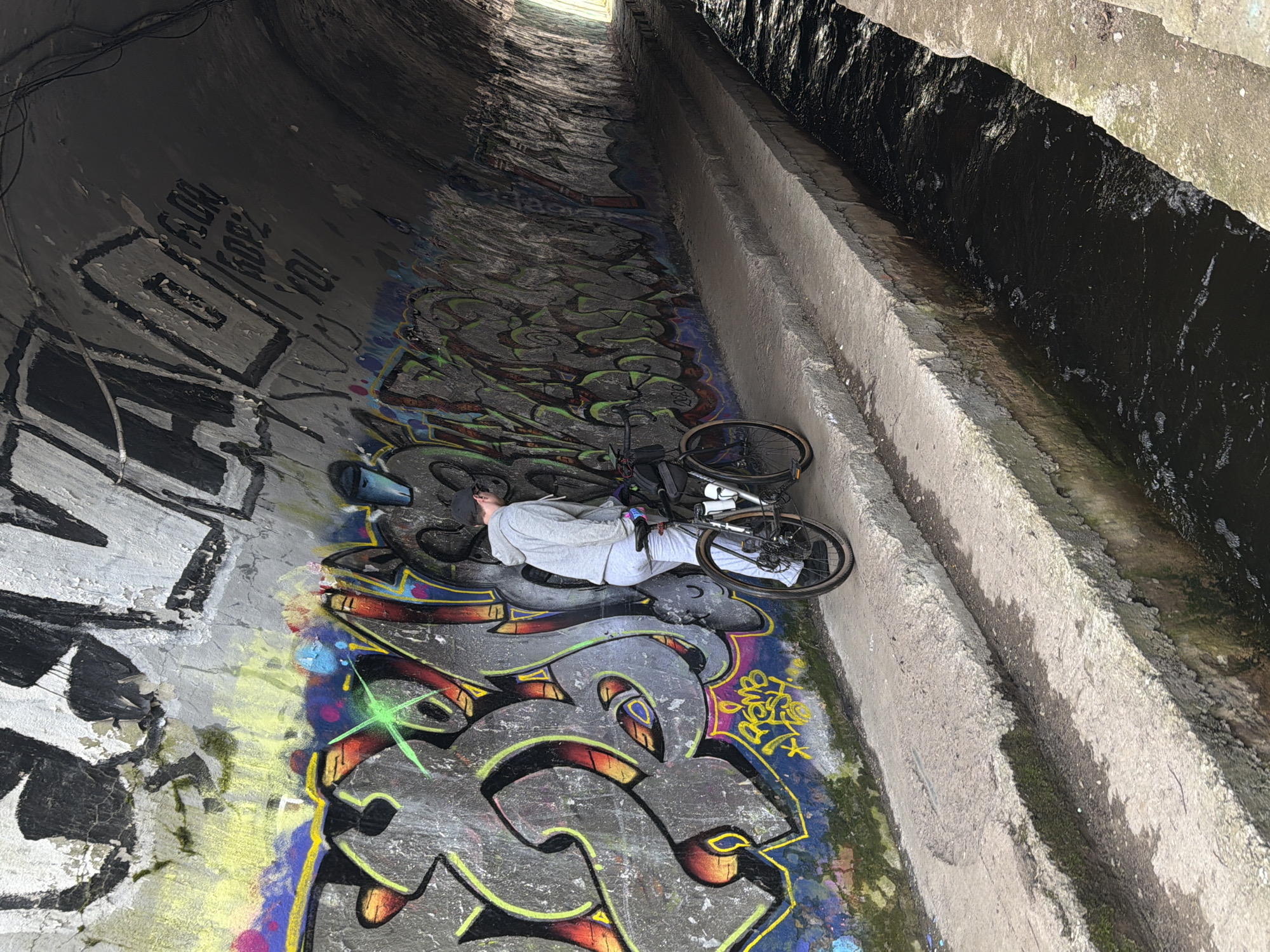A cyclist wearing light grey clothing lies on a graffiti-covered floor next to their bicycle in a concrete culvert. A narrow stream runs along the right side of the culvert. The culvert appears to be in a state of disrepair, and there is visible water damage and staining on the concrete. Based on the location provided, this scene is potentially located in Udelnaya, Russia.