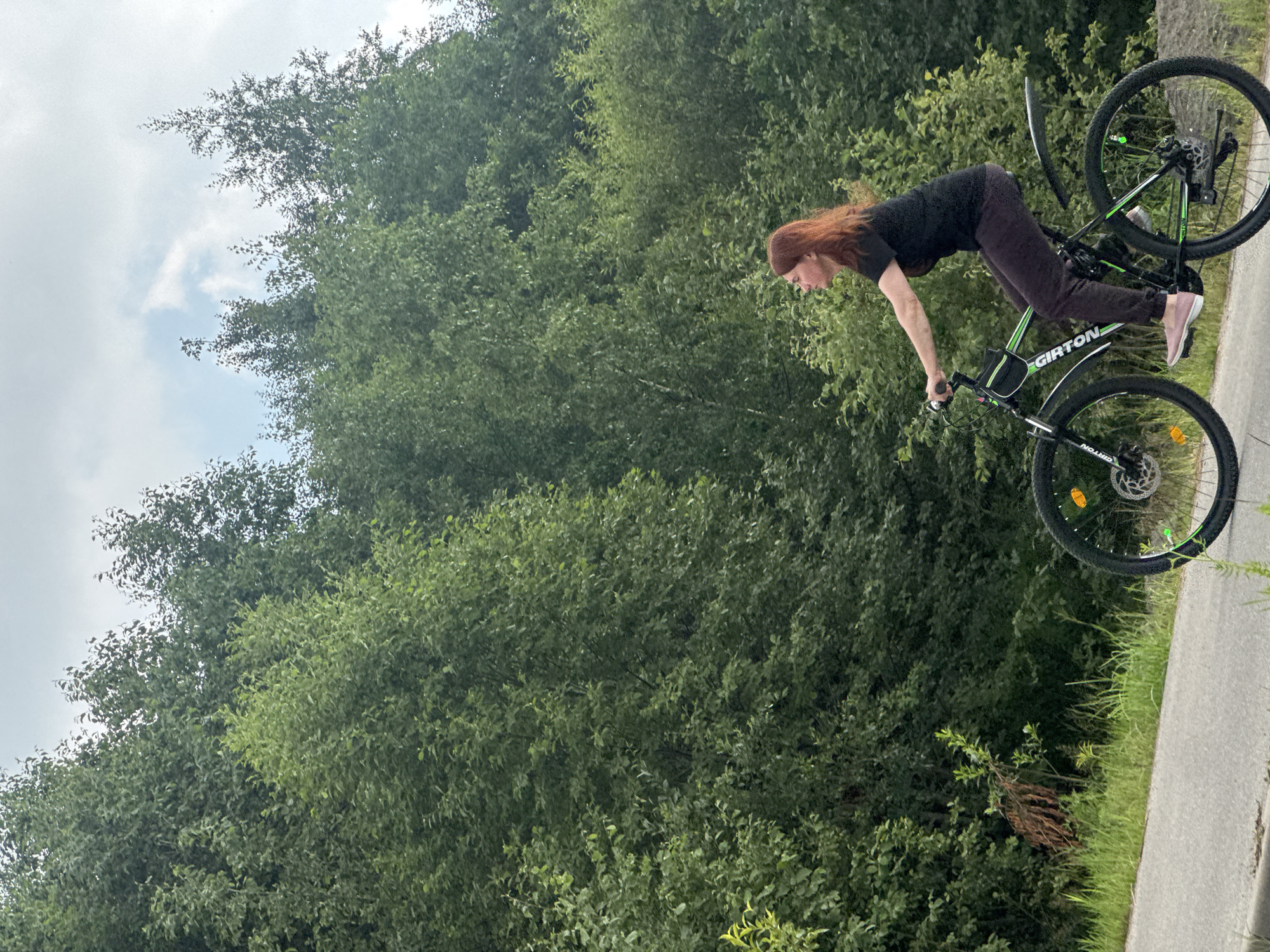 In Novoe Devyatkino, Russia, a woman with long, red hair is captured in a dynamic moment performing a stunt on a bicycle. She is wearing a black t-shirt and dark pants, with light pink shoes. The bicycle is predominantly black and green, with the brand name "GIRTON" visible on the frame. The woman is in mid-air, seemingly balancing on the rear wheel of the bike while leaning forward, her hands gripping the handlebars.
The scene is set against a backdrop of dense, lush green foliage, suggesting a park or natural area with abundant trees. The sky above is overcast with some patches of light, indicating a cloudy day. The ground appears to be a paved path or road, bordered by grass and more greenery. The time of day is difficult to determine precisely due to the lighting, but it seems to be daytime. There are no overt emotional cues, but the pose suggests focus and athleticism. The "GIRTON" text is clearly visible on the bike's frame.