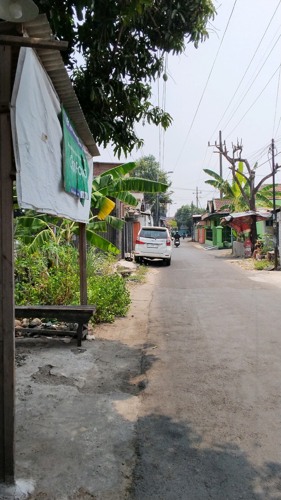 The image depicts a street scene in Banjaragung, Indonesia. In the foreground, a weathered wooden bench sits near a concrete area partially covered by shade. A tall, thin wooden pole supports a section of corrugated metal roofing and a white tarp with a green sign partially visible, displaying text in Indonesian. Lush green banana plants and other vegetation grow to the left of the bench.
The street itself is paved with asphalt and appears to be a residential or semi-rural road. A white minivan is parked on the side of the road, facing away from the camera. Further down the street, a single motorcycle with a rider is visible, moving away from the viewer. The surroundings are characterized by houses with colorful facades, predominantly green, and various trees, including a prominent mango tree with ripening fruit. Utility poles and electrical wires crisscross the sky. The overall lighting suggests it is daytime, with a bright, slightly hazy sky. The scene conveys a typical everyday atmosphere of a community in Indonesia.