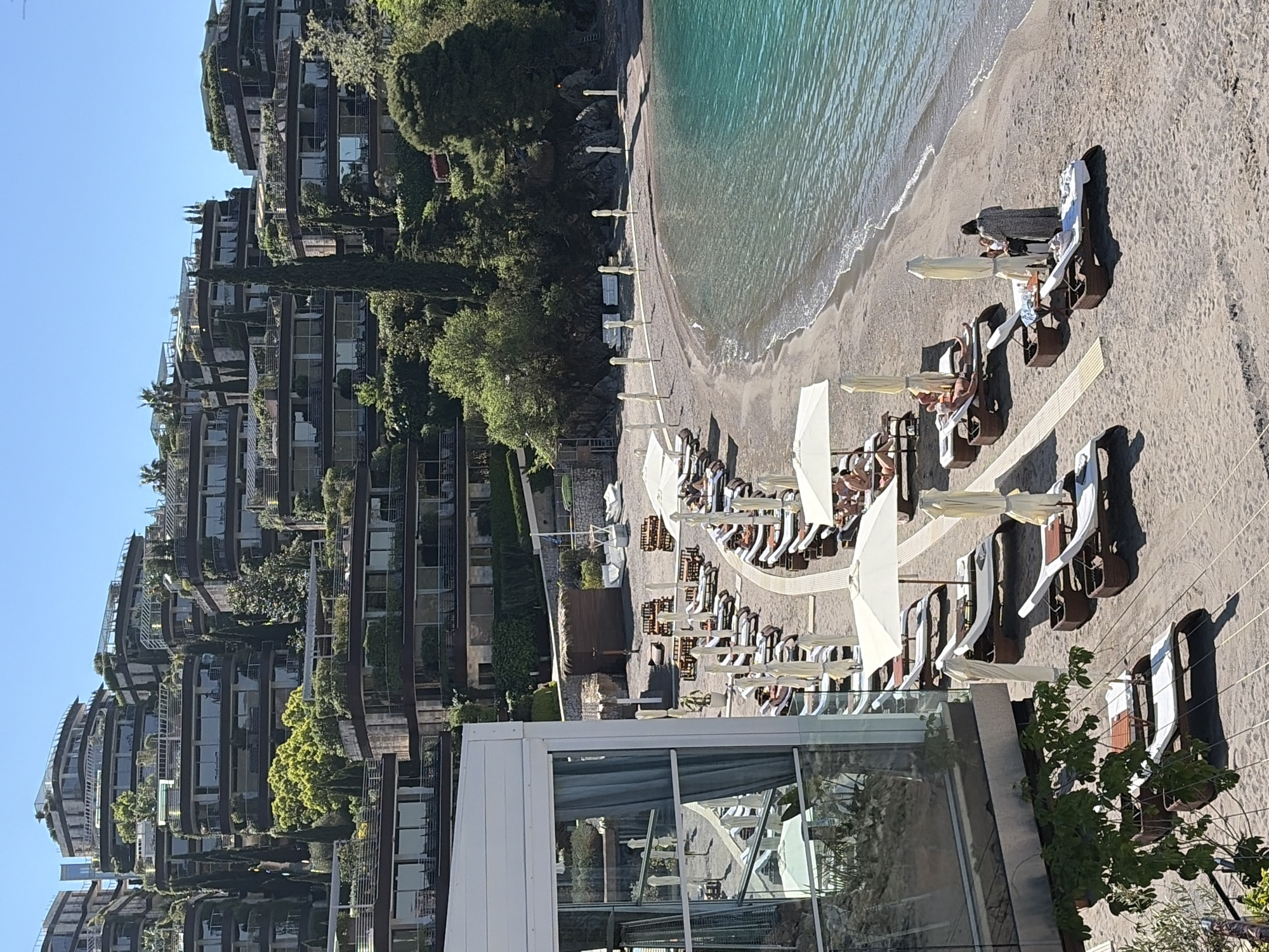 The image depicts a beach scene in Budva, Montenegro. A curved beach with turquoise water is lined with rows of white beach chairs and sun umbrellas. People are relaxing on the chairs. Next to the beach is a building with glass windows and a white exterior. Beyond the building, a hillside is covered with multi-story buildings with greenery on balconies. The sky is clear and blue.