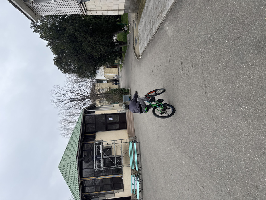 The image depicts an outdoor scene in Anapa, Russia, under an overcast grey sky. An asphalt paved area occupies the foreground and center of the frame. A single individual, appearing to be a child or young adolescent, is shown from the rear, riding a green bicycle with black tires and red accents. The individual wears a grey jacket, grey patterned trousers, and a grey helmet, and is positioned in the lower-center of the image, facing away from the viewer.
To the left, a multi-sided building with light beige walls and a green tiled roof is visible. This structure features numerous dark-framed windows and a brown double door entrance with steps. Metal scaffolding is erected against one section of windows, where a figure, presumably a worker, is present and appears to be actively working. Two light-blue metal benches are positioned on the asphalt in front of this building.
The background features a curved asphalt path extending into the distance, with several indistinct figures walking further along. On the right side of the path, a large, dark green coniferous tree stands adjacent to a light-colored brick building with shingle-style siding on its upper portion. A metal anti-tank barrier (hedgehog) is visible on the ground near the base of the tree. Bare-branched trees and additional light-colored buildings are present further in the background.