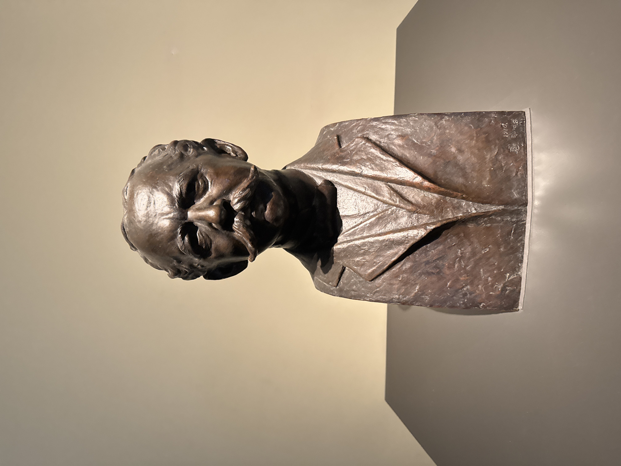 A bronze bust of a man with a prominent mustache stands on a dark grey pedestal against a light beige wall. The bust depicts the man from the shoulders up, wearing a suit jacket and a collared shirt with a tie. His hair is styled back, and he has a stern expression. There appears to be a signature and date inscribed on the right side of the bust. The lighting creates shadows on the wall behind the sculpture, suggesting it is indoors, likely in a museum or gallery setting in Bern, Switzerland.