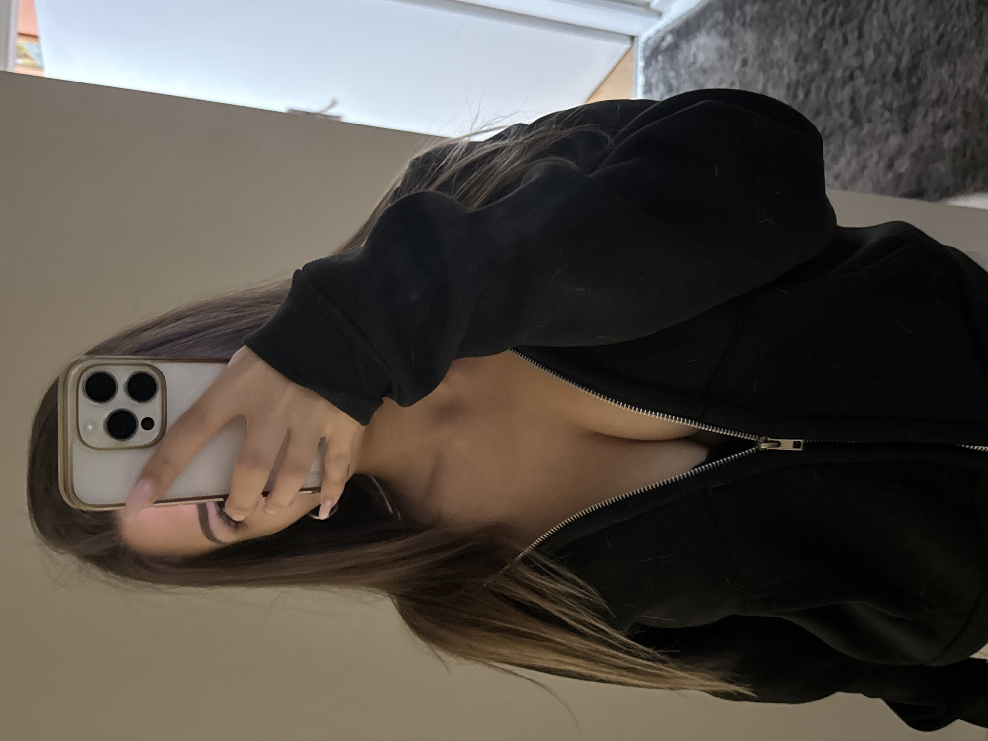 A woman with long brown hair, wearing a black zip-up hoodie, is taking a selfie in a room with beige walls. Her face is partially obscured by a white iPhone with a light-colored case. The background includes a doorway and a glimpse of a gray, textured rug or carpet. The location of Sofia, Bulgaria cannot be determined from the image.