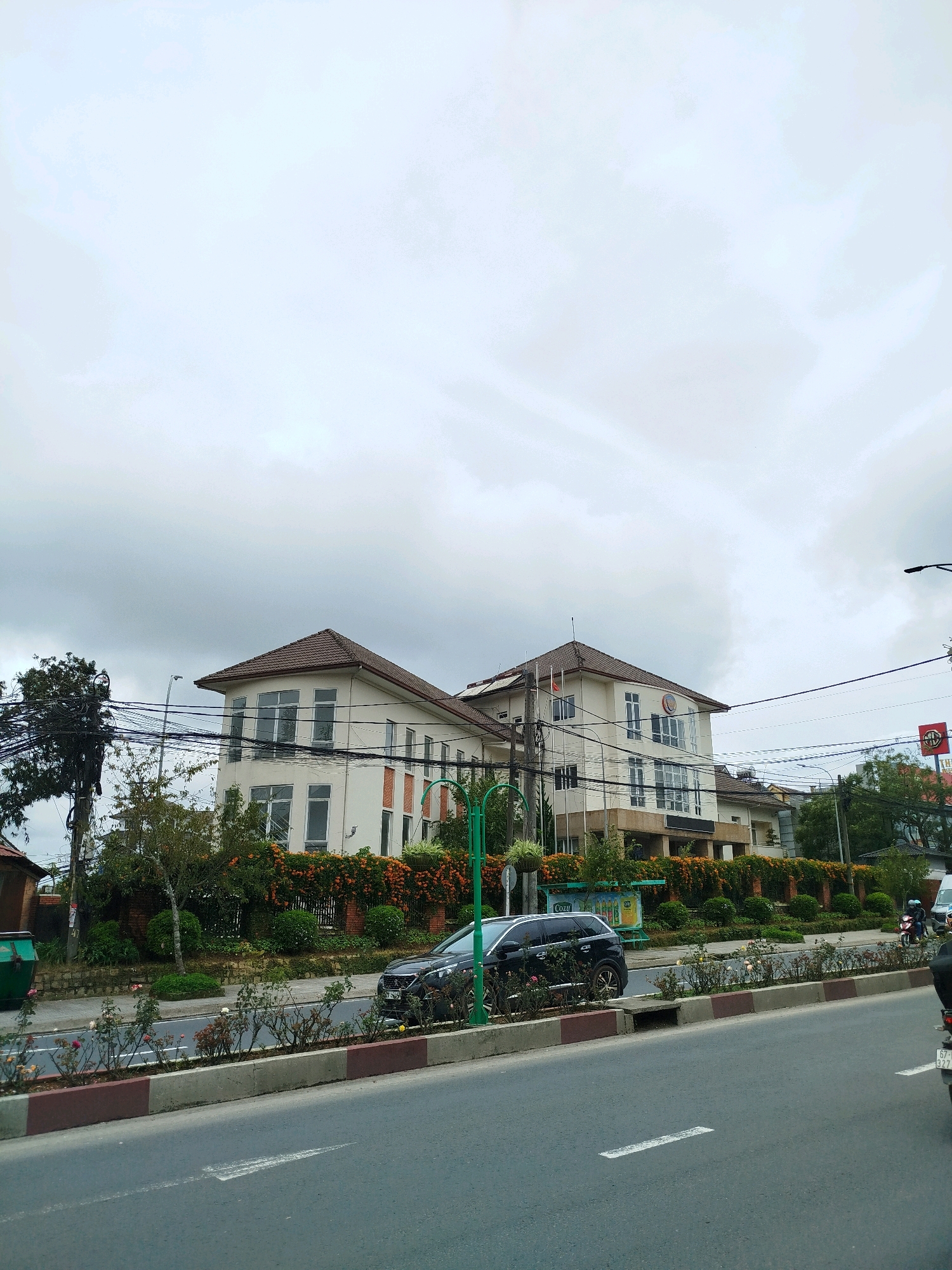This is a street-level, medium-shot photograph taken in Da Lat, Vietnam, under an overcast sky. The main subject is a large, cream-colored building with a brown tiled roof, featuring multiple stories, numerous windows, and architectural details such as decorative brickwork. The building appears to be a commercial or institutional property, with a visible logo on its facade.
In the foreground, a paved road with lane markings is visible, suggesting a moderately busy street. Several vehicles are present, including a dark-colored SUV parked along the roadside and a motorcycle with a rider further down the street. The roadside is lined with a decorative border and a planted median strip featuring rose bushes and other greenery.
Behind the building, a dense line of trees and bushes forms a natural barrier, with some flowering plants visible. A prominent utility pole with a complex network of electrical wires stands to the left of the frame, indicating urban infrastructure. A green dumpster is situated near the sidewalk. The overall environment suggests a well-maintained urban area, possibly a commercial district or a street with significant buildings. The weather appears to be cloudy and possibly humid.