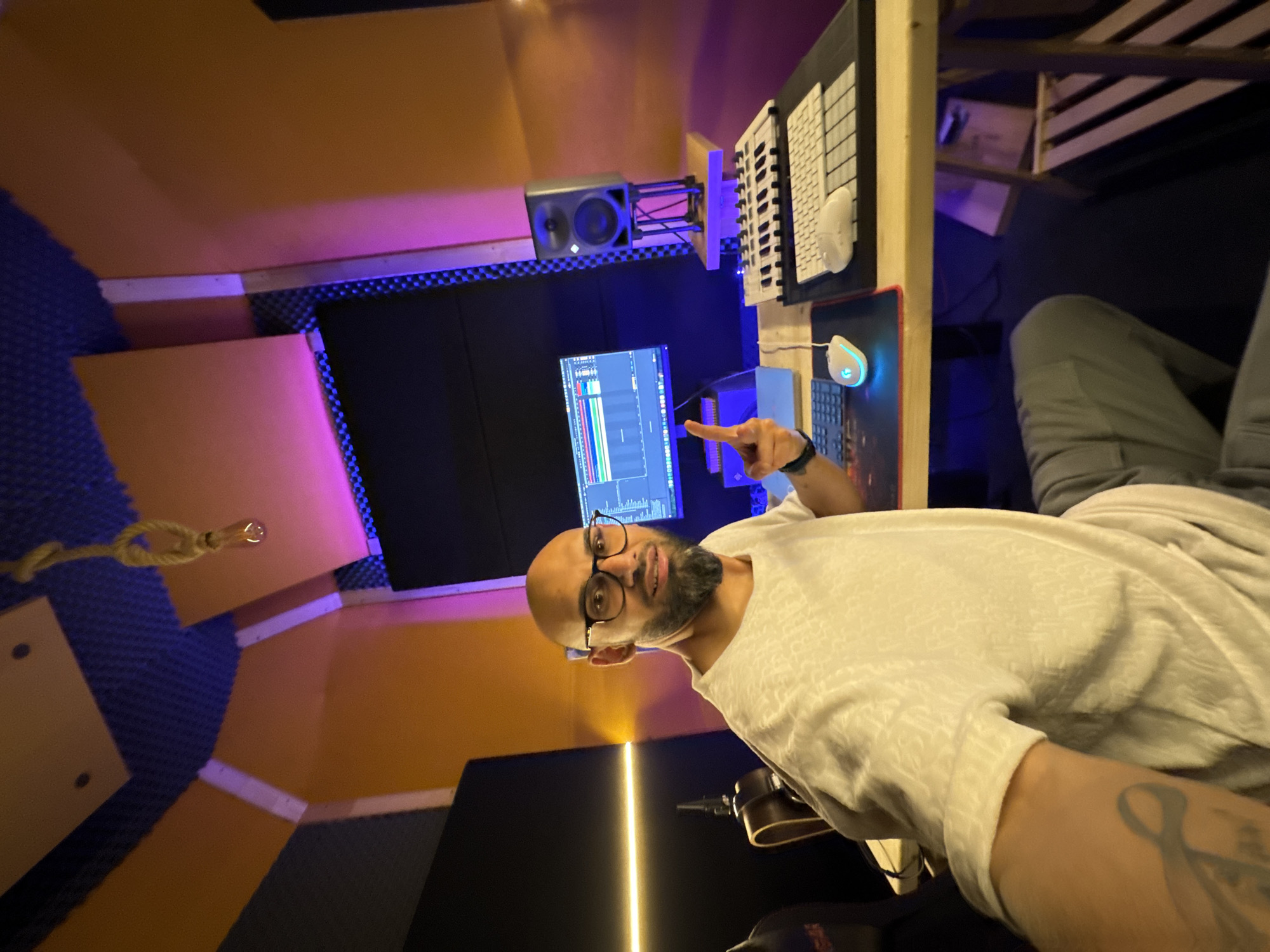 The image shows a man in a recording studio. He is bald with a beard, wearing glasses and a white t-shirt. He is pointing towards a computer monitor displaying audio editing software. The studio features soundproofing panels in various colors and blue LED lighting. Equipment includes a keyboard, a monitor speaker, and a mouse. The studio is likely located in Plovdiv, Bulgaria.