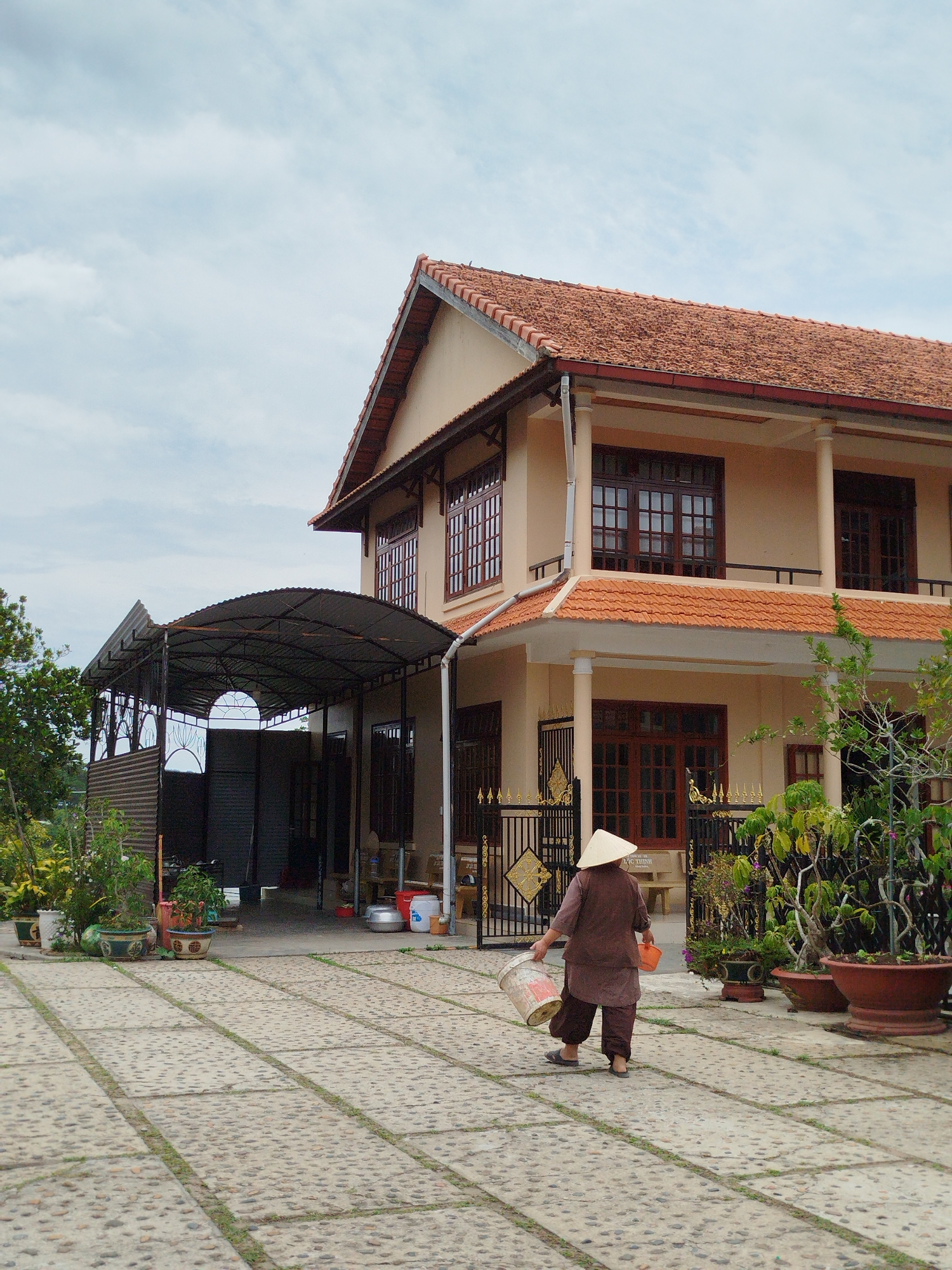 A medium-shot captures a person walking away from the viewer, carrying a bucket in one hand and a smaller orange container in the other, on a paved surface in front of a two-story building. The person wears traditional Vietnamese clothing in brown hues and a conical hat. The building has a terracotta tiled roof, beige walls, dark brown window frames, and decorative iron railings. Potted plants line the front of the building. A metal carport structure extends from the side of the building. The paved area is made of stone blocks. The sky is overcast. The location is Da Lat, Vietnam.