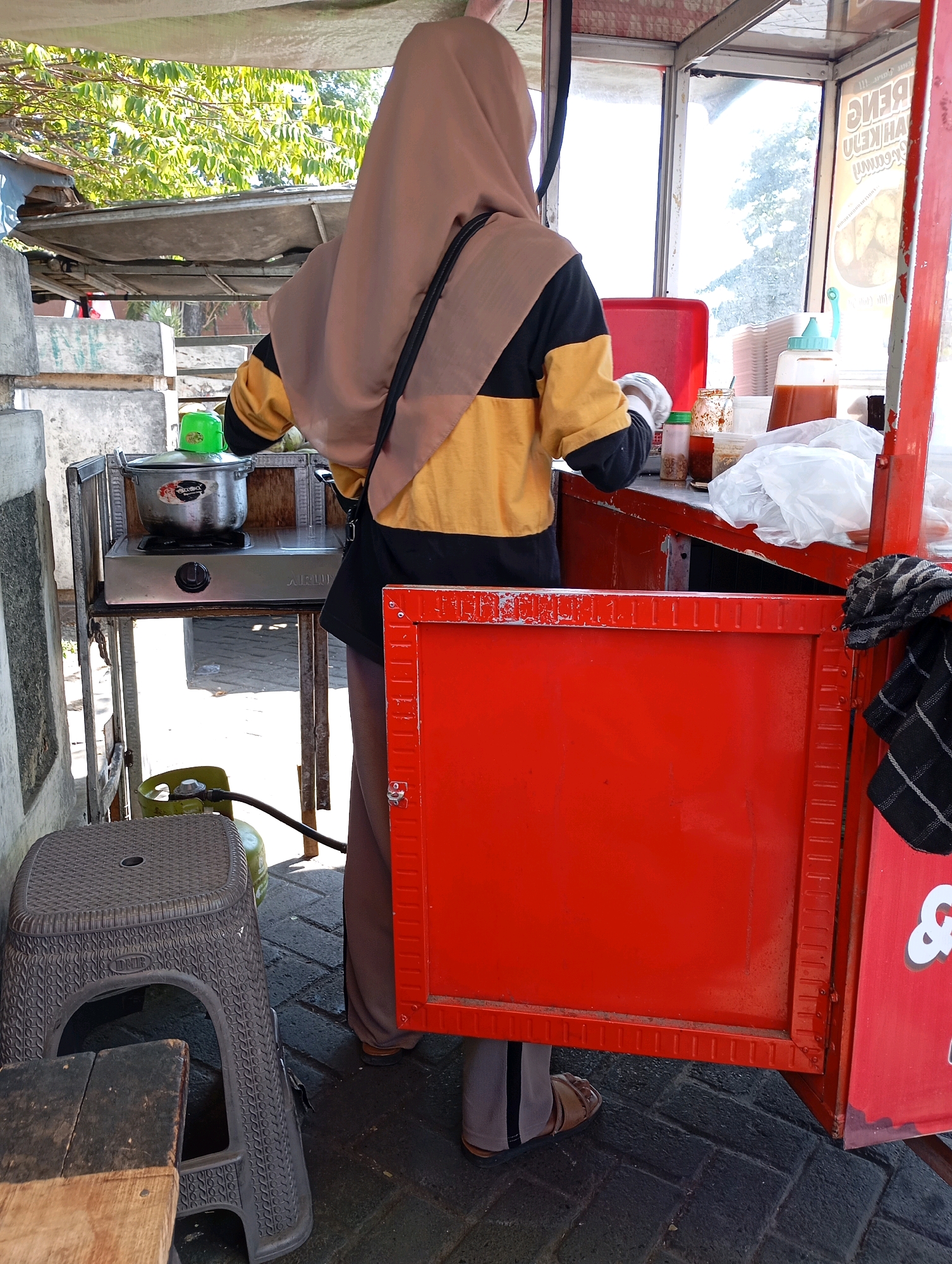 In the city of Kalisampurno, Indonesia, a person wearing a hijab and a yellow and black long-sleeved shirt stands behind a food cart. The cart has a red front panel and a sign with text that appears to be in Indonesian. To the left of the cart, a gas cylinder is connected to a portable stove, on which a pot with a green lid is placed. A grey plastic stool and a wooden bench are visible near the cart. The ground is paved with dark stones. The background shows trees and a bright sky, indicating an outdoor setting. The person is preparing food, with various containers and utensils on the counter of the cart.