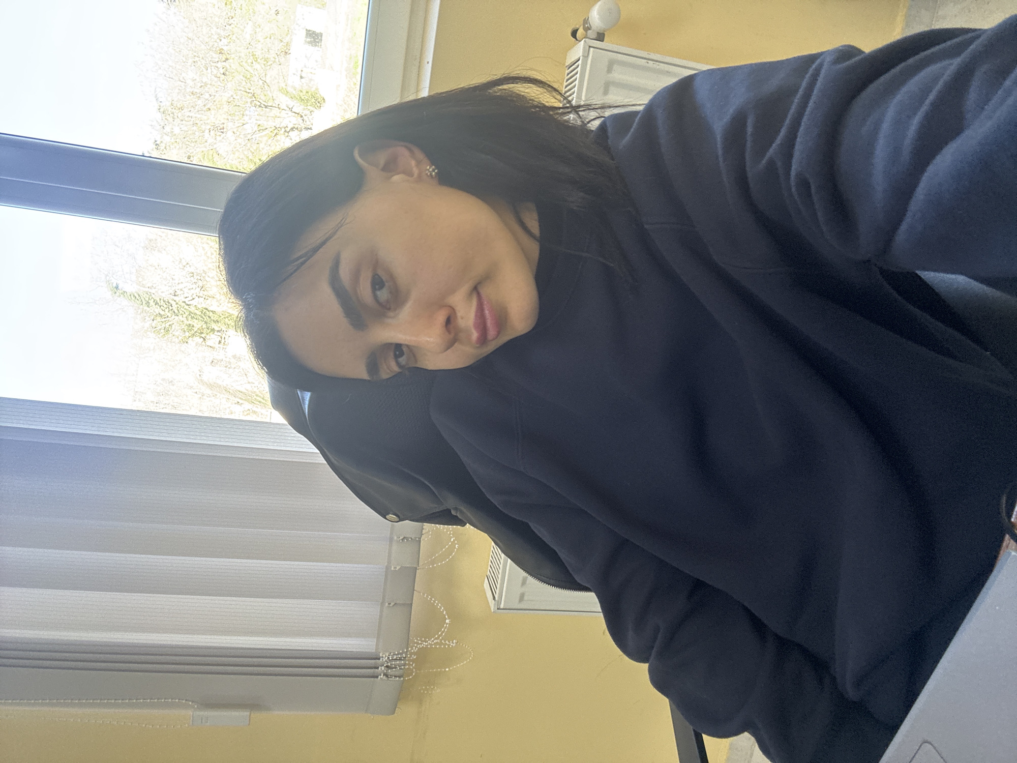 A young woman with dark hair and fair skin is positioned at an angle, partially resting her head on her arm. She wears a dark blue sweatshirt. The background includes a window with blinds, an outdoor scene with trees, a yellow wall, and a radiator. Based on the information given, the location is Tbilisi, Georgia. A laptop is visible on a table.