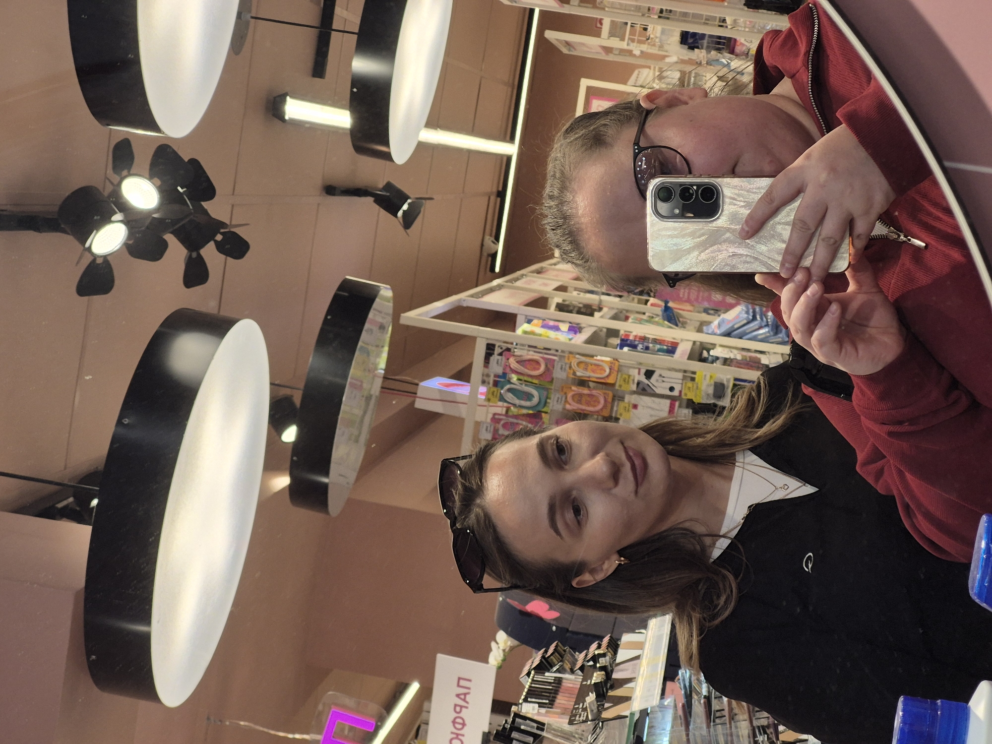 A mirror selfie in a store in Chaikovsky, Russia. Two women are reflected in the mirror. The woman closer to the foreground has brown hair, sunglasses on top of her head, and is wearing a black jacket. The second woman is wearing a red jacket and eyeglasses, and is holding a smartphone, presumably to take the selfie. The store appears to sell cosmetics and toiletries. The word "ПАРФЮМ" is visible in Cyrillic on a product display. Circular light fixtures hang from the ceiling.