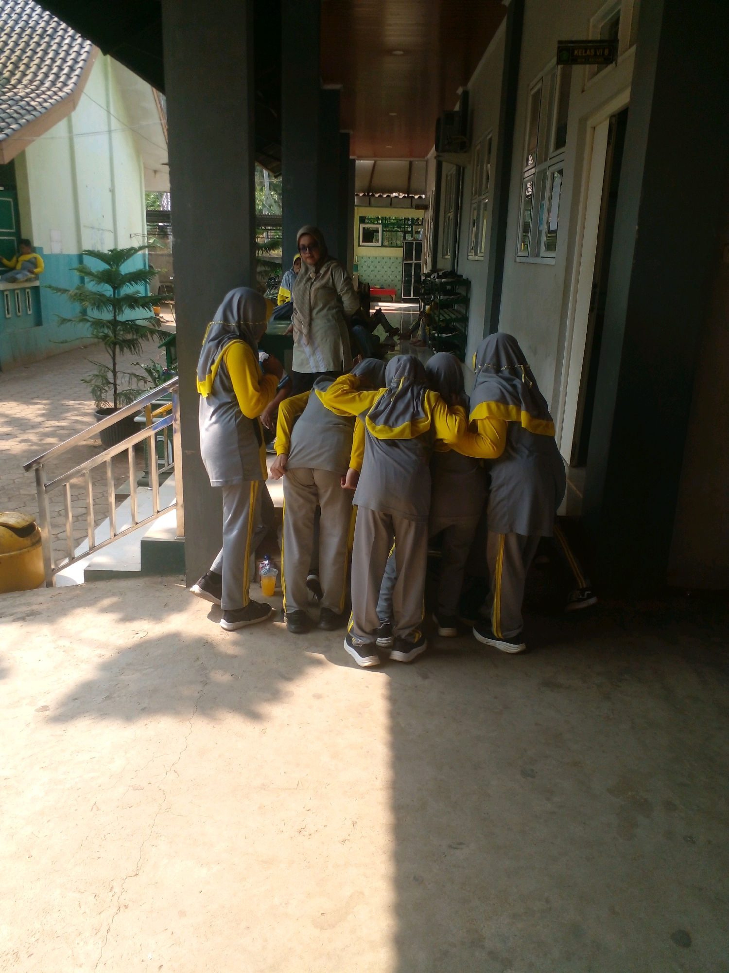 A group of six young people wearing gray and yellow uniforms, including hijabs, are huddled together in a circle, with their backs to the camera. They appear to be students. A woman in a beige top and glasses stands to the side, looking at the students. They are on a paved walkway outside a building with a gray pillar and a brown roof. There is a metal railing with stairs to the left, and a yellow trash can. In the background, there is a light green building with a dark green door and windows, and a small pine tree. The scene appears to be in a school or educational institution located in Banyuputih, Indonesia.