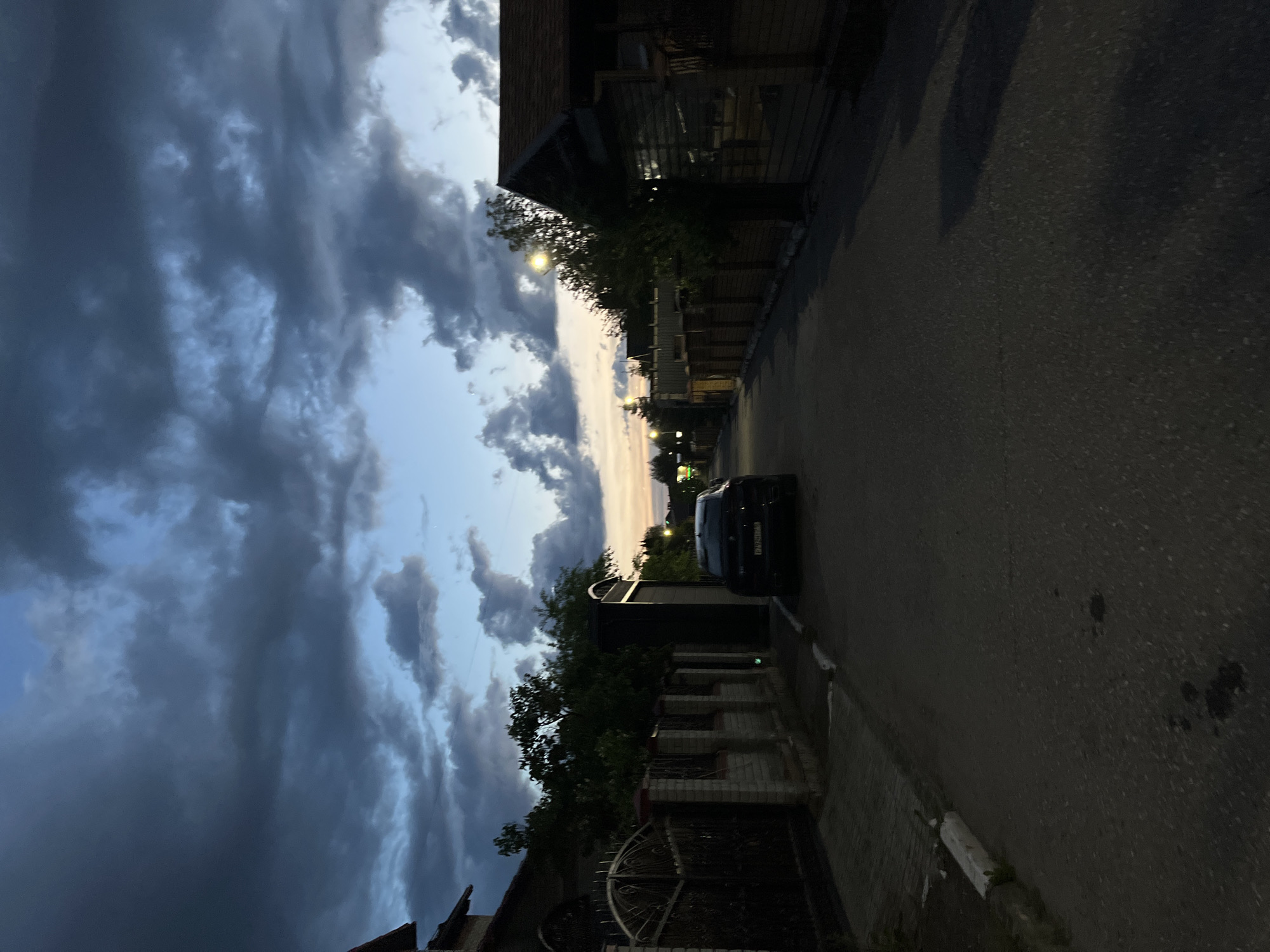 The image captures a quiet street scene at dusk or early night in Ореховка, Russia. The sky is largely covered by dark, heavy clouds, but a lighter band of sky with faint yellowish and blue hues is visible towards the horizon, indicating twilight. Below the sky, a street stretches into the distance, lined with dark-colored buildings and green trees. Streetlights cast warm glows, providing artificial illumination to the otherwise dim environment. A dark-colored car, likely a sedan or SUV, is parked on the right side of the street, facing away from the viewer. Its license plate, `К176УР 197`, is partially visible. On the left side of the street, a decorative, possibly wrought-iron, gate and a brick fence are discernible. No people or overt activities are visible, lending a peaceful and somewhat somber atmosphere to the scene. The low light and cloud cover suggest either a recent rain or simply a heavily overcast evening.