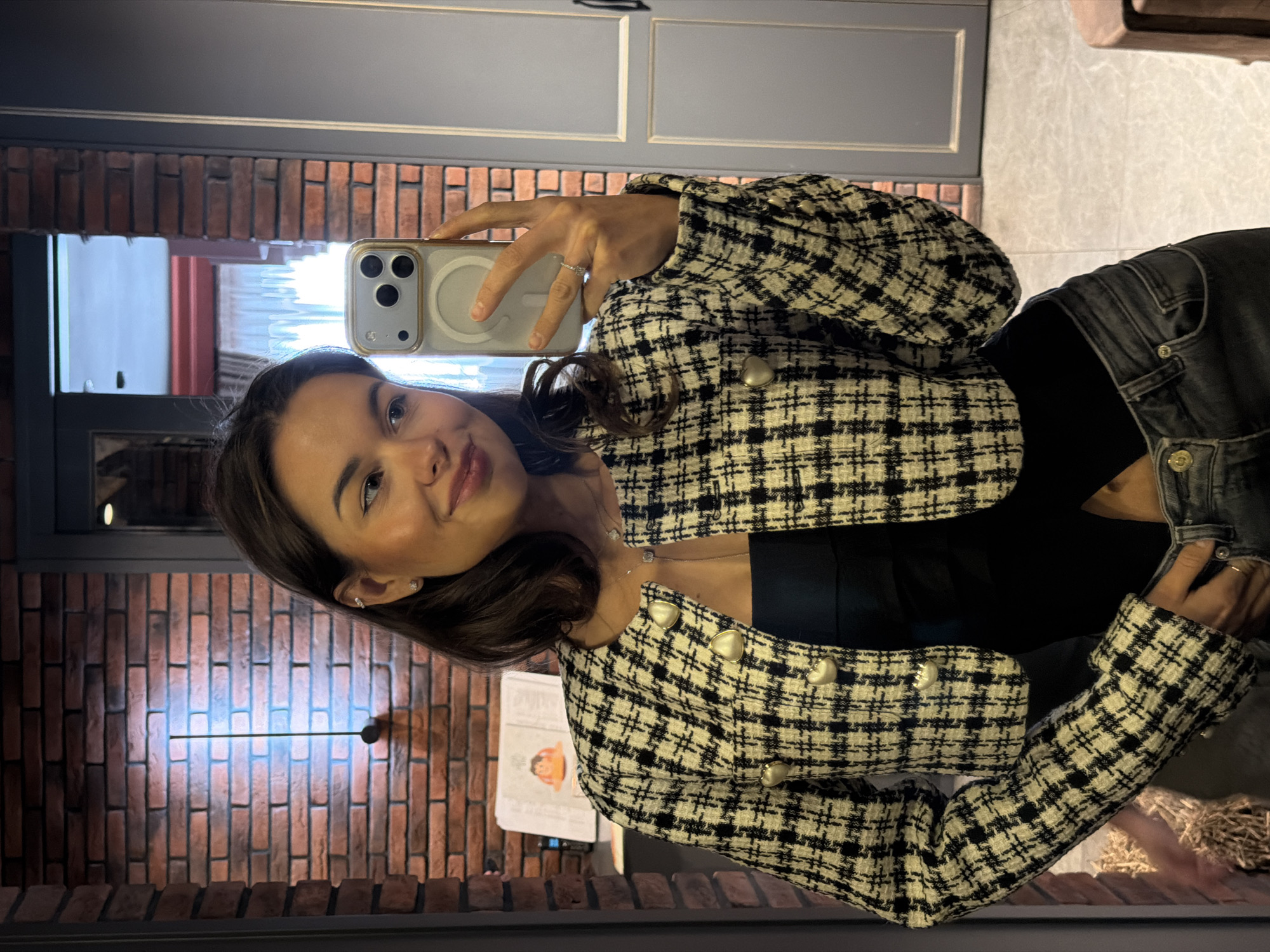 A woman is taking a selfie in front of a mirror. She is wearing a black and white tweed jacket with gold heart-shaped buttons, a black top, and dark-wash jeans. The woman is holding her phone in her right hand. The background consists of a brick wall with a horizontal linear light fixture, a light gray painted wall, and a door. A book is on a shelf. The location appears to be an interior space, possibly a private residence, in Krasnoyarsk, Russia.
