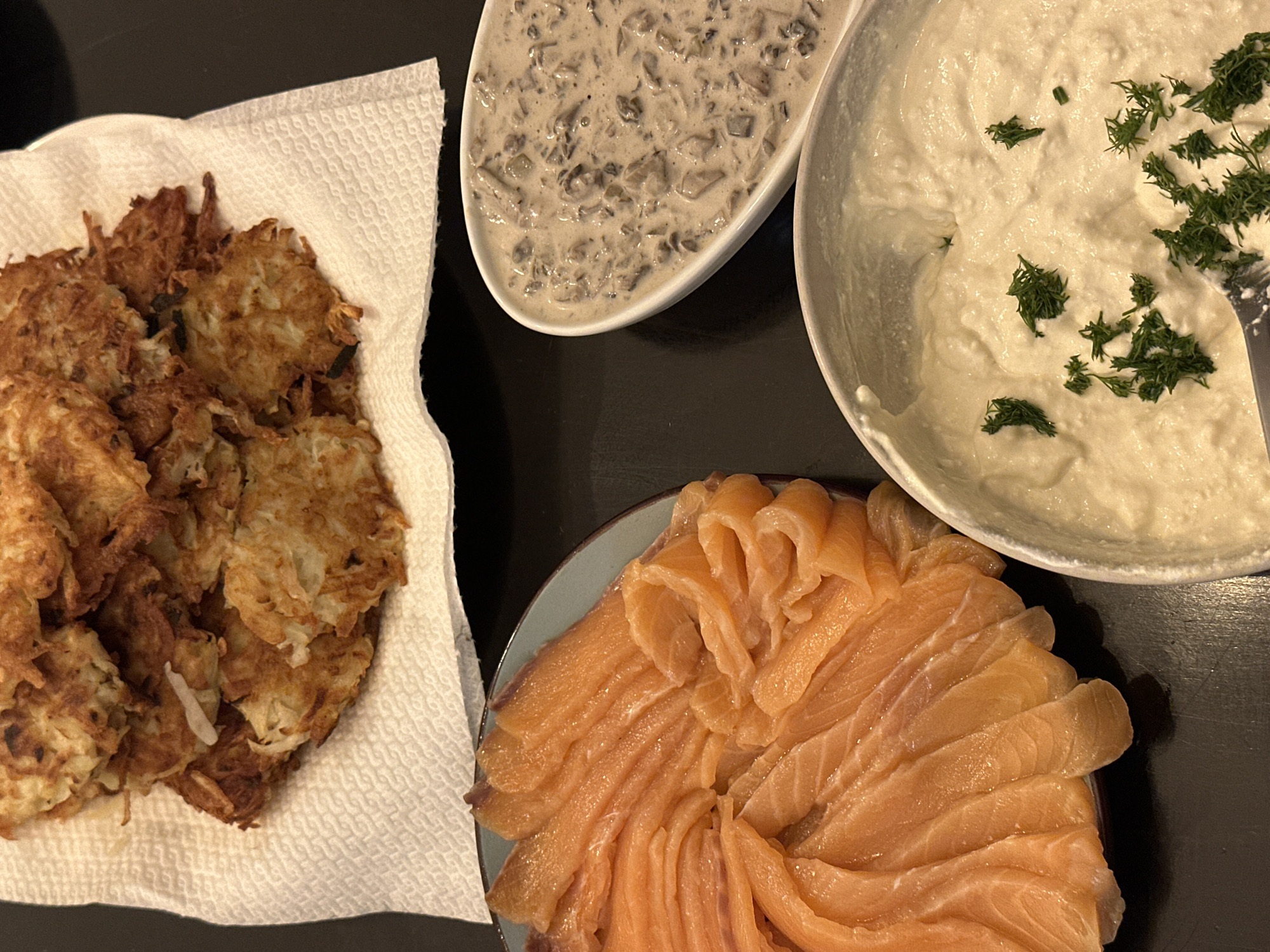 The image shows a table with four dishes: a stack of potato pancakes on a white paper towel-lined plate, a boat-shaped dish of mushroom sauce, a bowl of sour cream topped with dill, and a plate of sliced salmon. The dishes are arranged on a dark surface. The location in the city of Ramat Gan, Israel, is not discernible from the image.