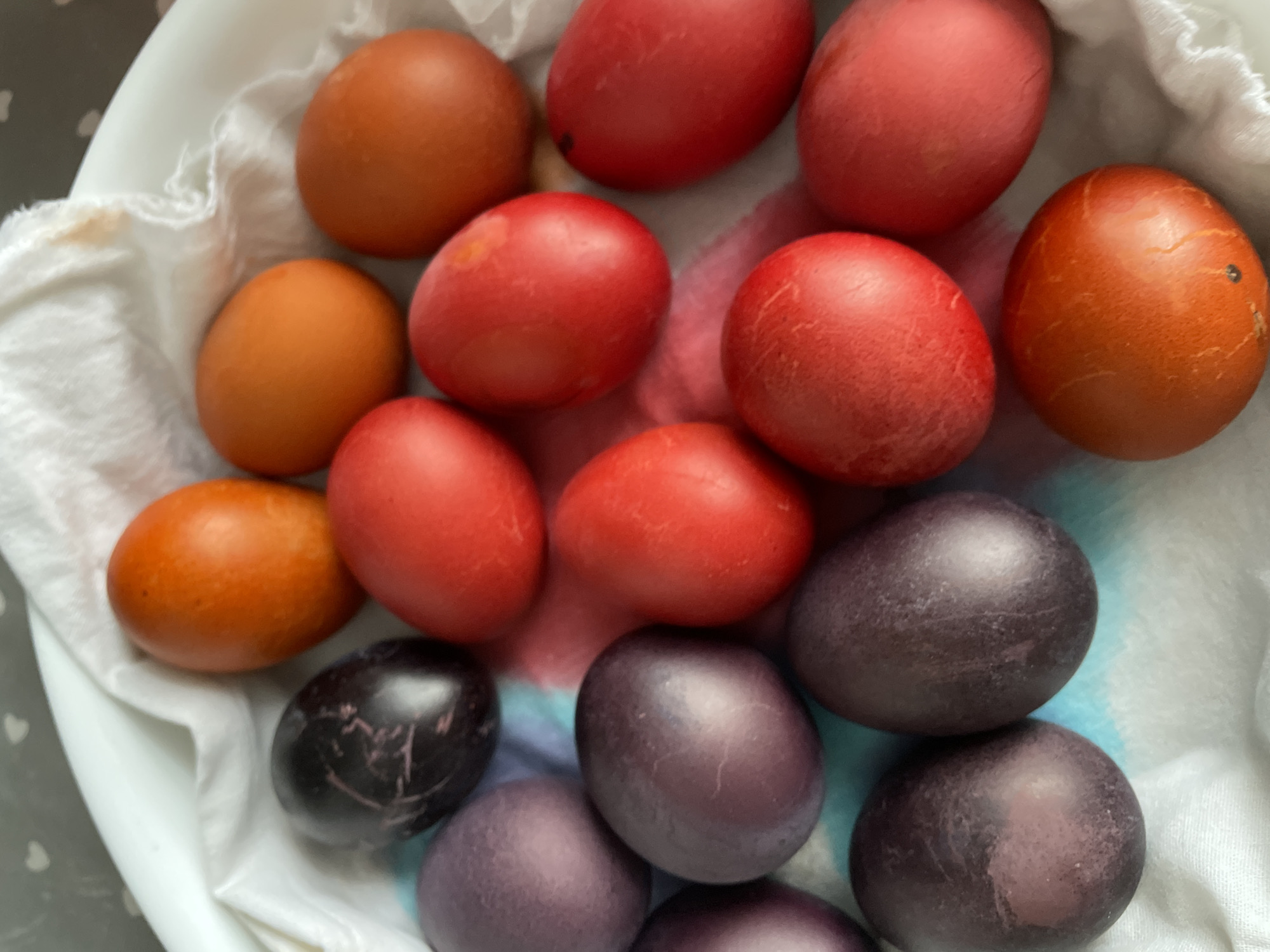 A close-up overhead shot shows a collection of Easter eggs of varying colors arranged on a white cloth inside a white bowl. The eggs are dyed in shades of brown, red, and purple/black. The colors seem to be naturally derived. Some of the eggs have blemishes or streaks in their coloring. The white cloth appears to be slightly damp. The bowl is positioned against a surface with a gray and white pattern. The location is not identifiable from the image.