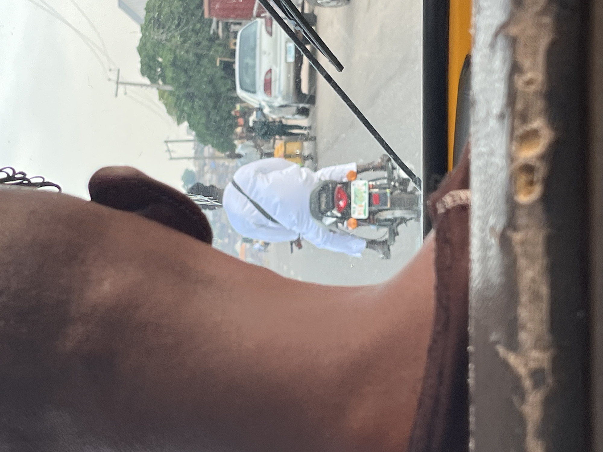 A person is seen from behind, wearing a white robe and a black sash, riding a motorcycle. The motorcycle has a license plate. In the background, other vehicles and people are visible on a road. The scene appears to be in a city. The image is taken from inside another vehicle, looking through a windshield. The focus is on the motorcycle rider and their attire.