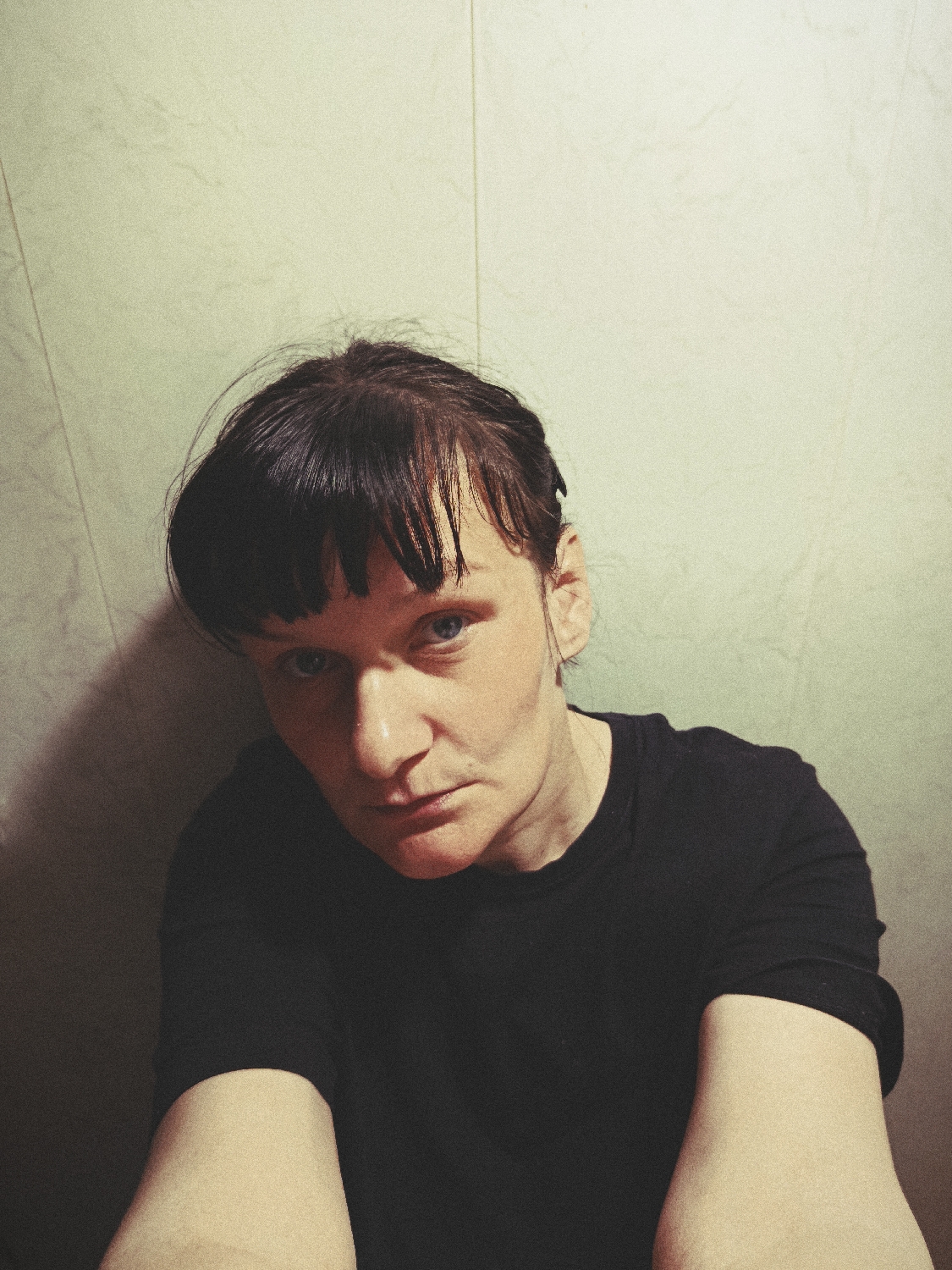 A close-up, chest-up portrait features an individual with short, dark hair and bangs, looking directly into the camera. The person has blue eyes and a serious, unsmiling expression. They are wearing a plain black t-shirt. The lighting is dramatic, with the right side of the face and part of the upper body illuminated more brightly, while the left side is in shadow. A distinct shadow is cast on the light-colored, textured wall behind them, suggesting a light source from the right. The background wall appears to be a light off-white or very pale green, with subtle vertical lines. The image has a grainy texture, possibly due to low light conditions or an applied filter, giving it a somewhat retro or filmic aesthetic. No specific objects or visible text are present. The setting appears to be indoors, likely against a plain wall. The individual's pose suggests a self-portrait or a very close-up photo.