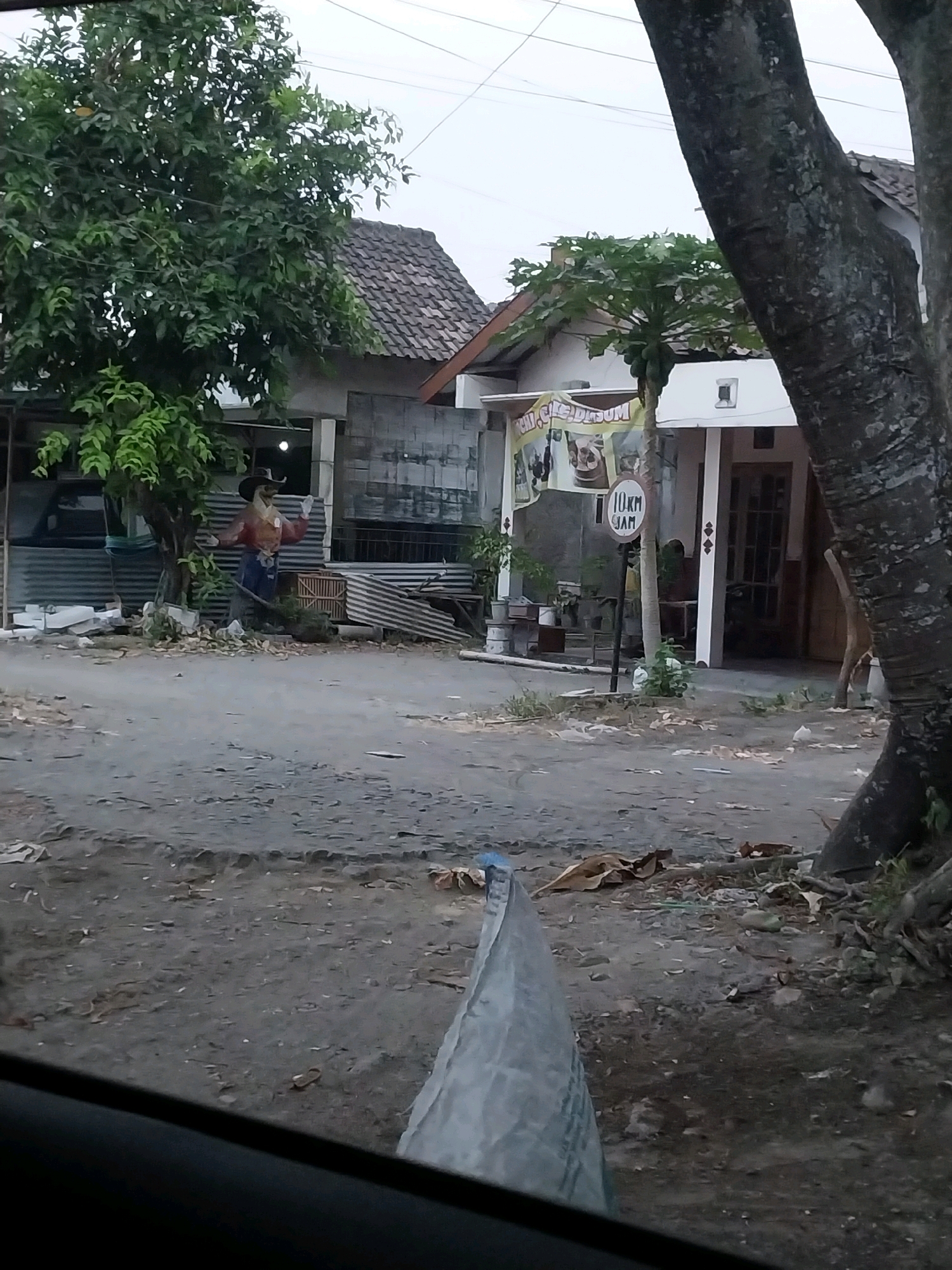 The image depicts a street scene in Maguwoharjo, Indonesia. A scarecrow dressed in a cowboy-like outfit stands near a tree on the left side of the frame. Behind the scarecrow, there is a building with a tiled roof and corrugated metal siding. A banner with text, partially obscured, is visible hanging on the front of a building to the right of the scarecrow. A circular sign indicating "10 KM JAM" is mounted on a pole in front of this building. The ground is unpaved and appears to be a mixture of dirt and gravel, with scattered debris. Trees, including a large one on the right, are present, and the sky is overcast. A portion of a vehicle's interior is visible in the foreground, suggesting the image was taken from inside a car.