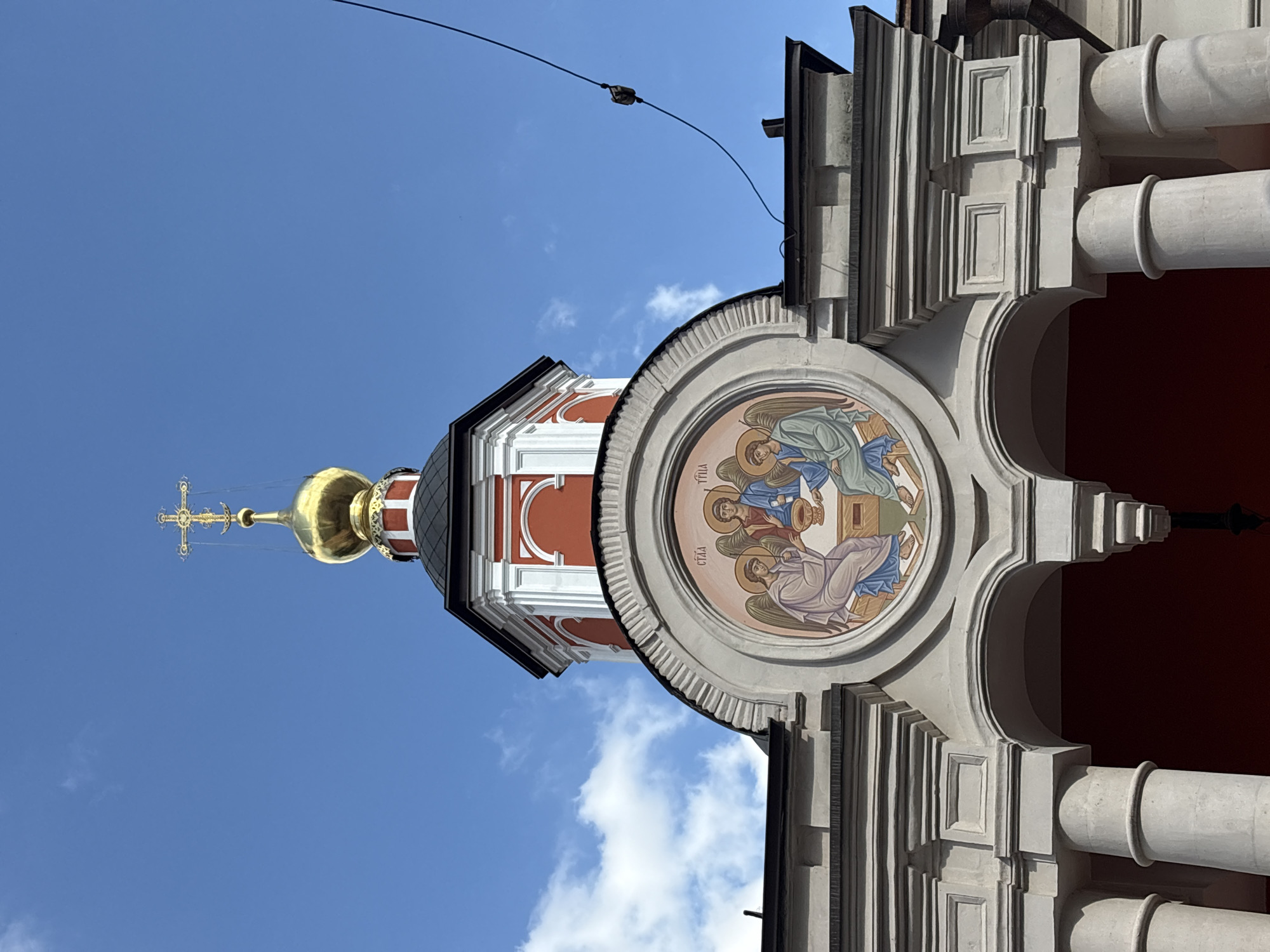 The image shows the facade of a building, likely a church, against a bright blue sky with scattered clouds. The architecture features ornate details, including columns and decorative moldings. A prominent circular fresco adorns the building, depicting three figures with halos and wings, suggesting a religious theme, possibly of angels. One figure appears to be holding a chalice. The building itself is painted in red and white, with a dark roof. To the left of the fresco and partially visible is a golden dome topped with a cross, characteristic of Eastern Orthodox churches. The lighting suggests it is daytime, likely mid-morning or afternoon, with clear weather. There are some faint Greek inscriptions visible on the fresco, but they are not clearly legible. A thin black cable stretches across the sky at the top of the frame. The image is oriented vertically, suggesting it was taken with a phone.